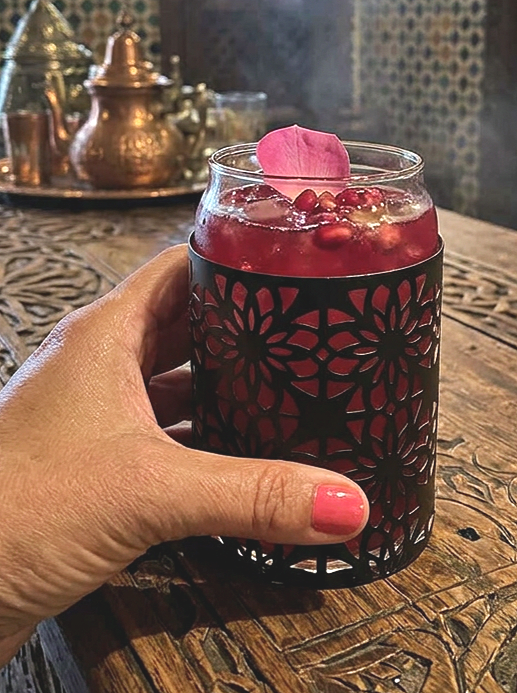 Moroccan inspired lattice cocktail glass serving a rose and pomegrante cocktail sitting on a bar top in a moroccan style bar and being held in a hand