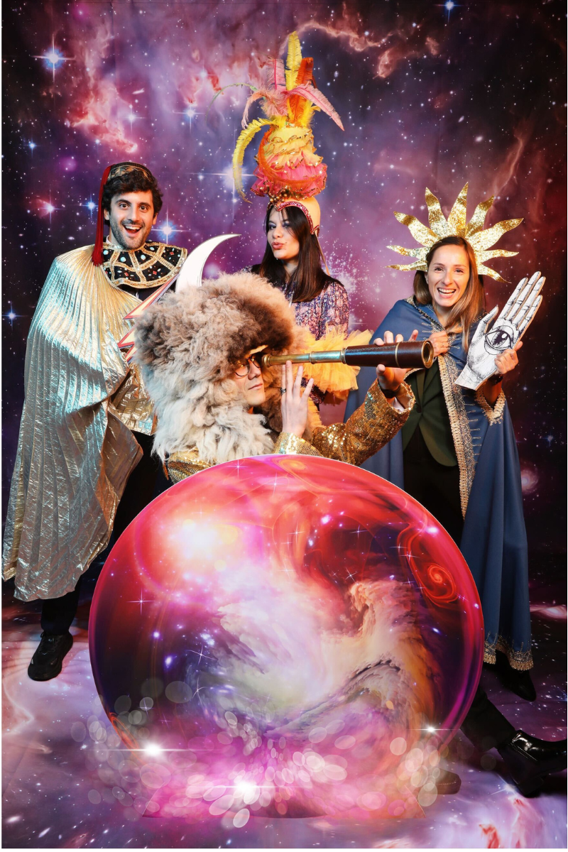Four people dressed in colorful, theatrical costumes posing in front of a cosmic backdrop with stars and nebulae. One person wears a fluffy white hat with horns, looking through a telescope. Another sports a large, vibrant feathered headpiece, and another has a gold star-shaped headdress. One person holds a cutout of an eye on their hand, and there is a large, swirling cosmic sphere in the foreground.