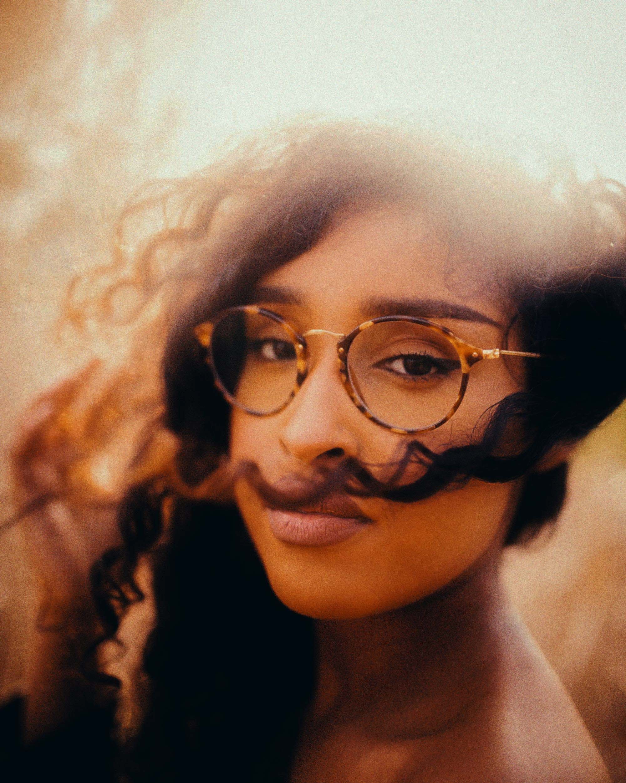 Close-up of a woman with curly hair wearing round tortoiseshell glasses, looking slightly away from the camera.