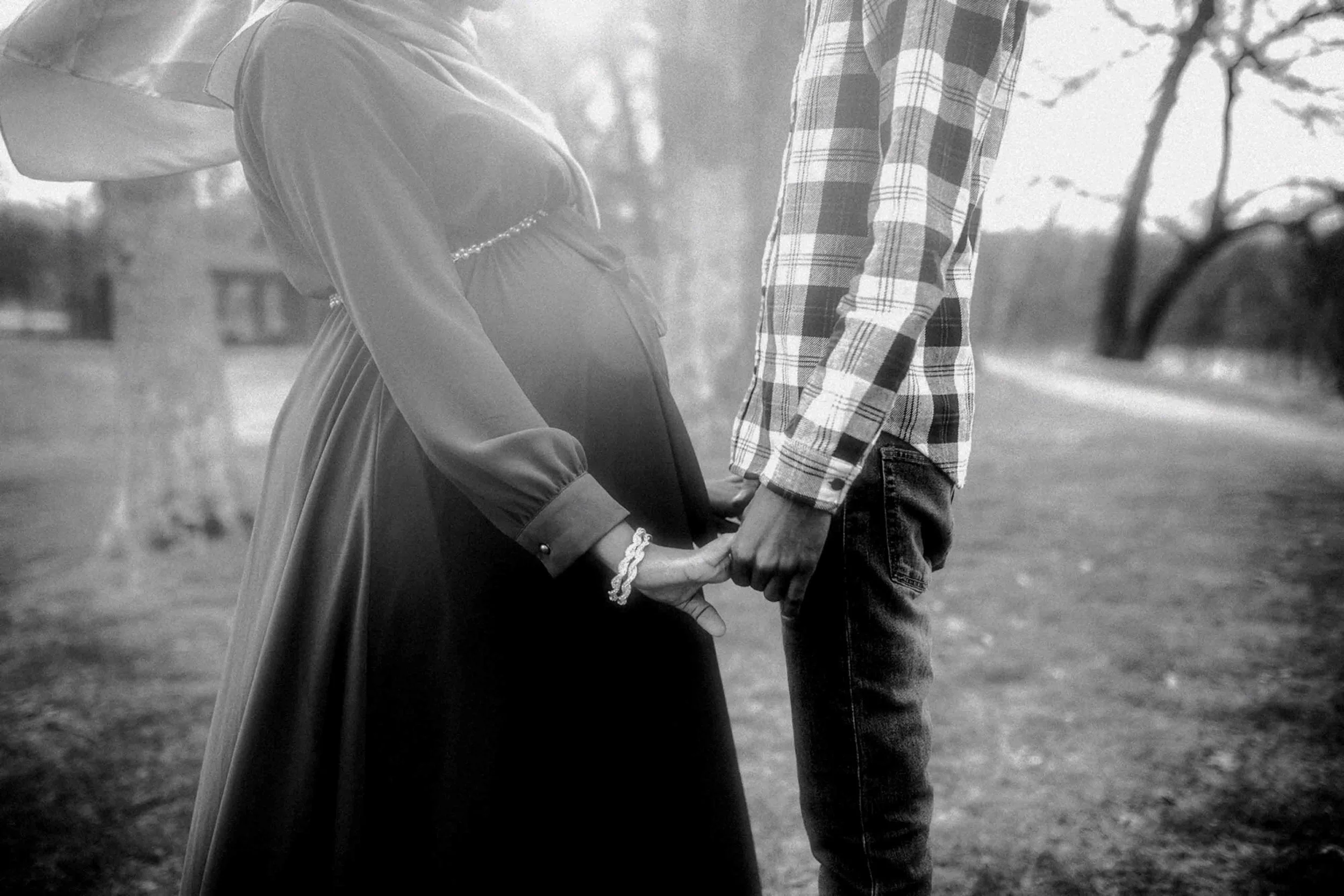A black-and-white photo of a pregnant woman and a man holding hands outdoors, standing on a dirt path with trees in the background.