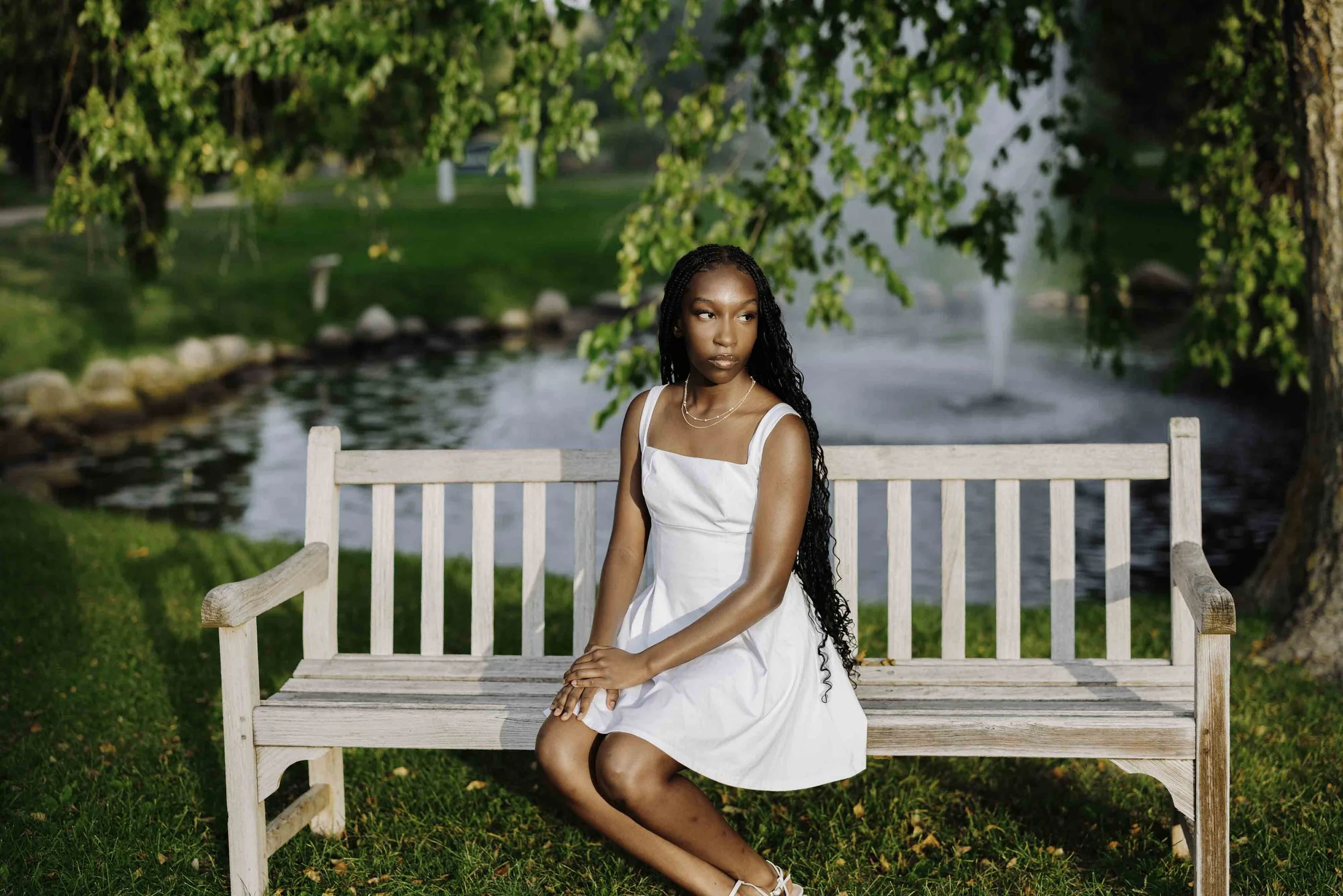 A young woman with long black curly hair, wearing a white dress, sitting on a wooden bench near a pond surrounded by green trees and grass, with a fountain in the background.