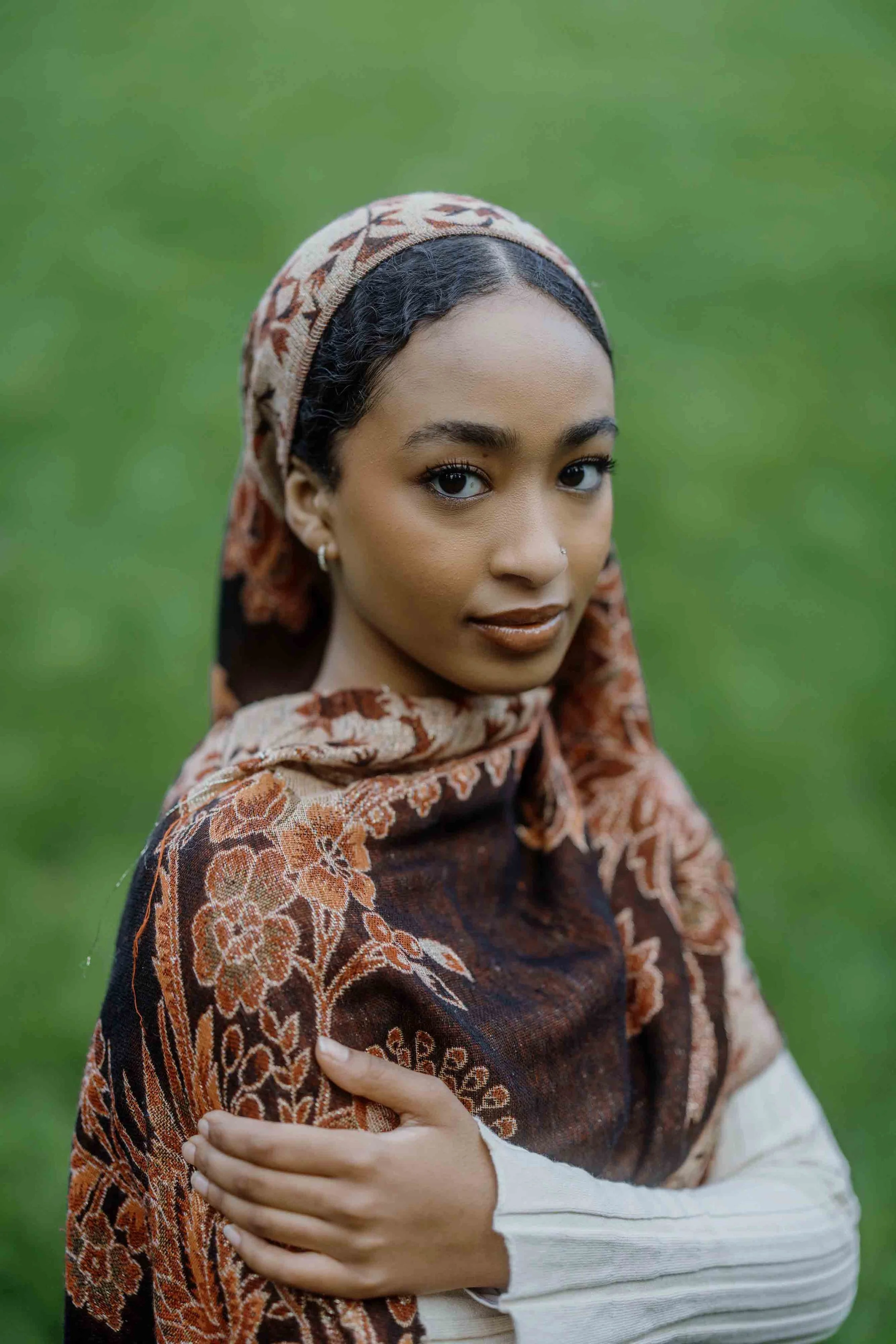 Portrait of a young woman with dark curly hair, wearing a colorful scarf with floral patterns, standing outdoors with a green blurred background.