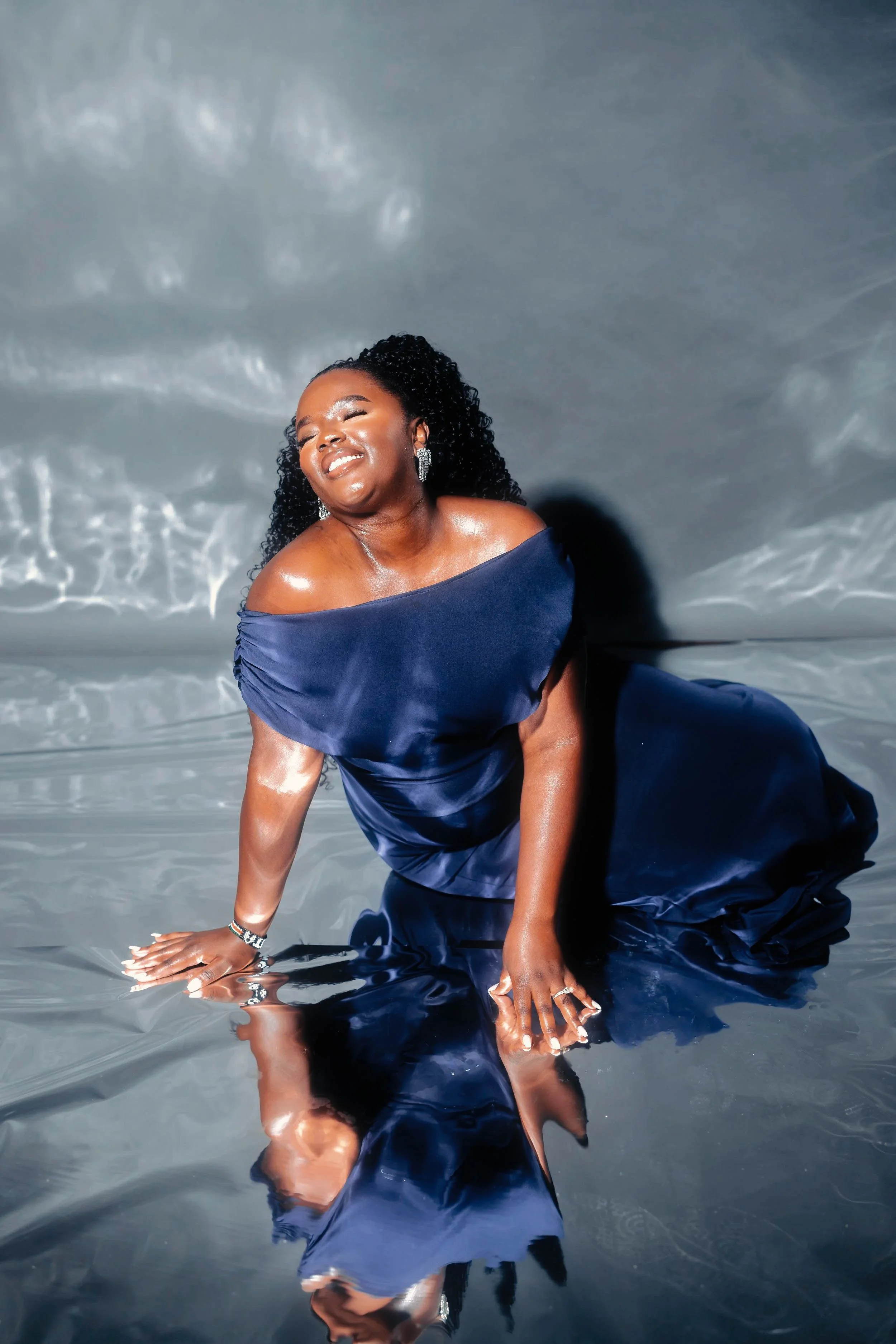 A woman with curly black hair, dressed in a navy blue satin gown, smiling and kneeling in a reflective water surface, with her reflection visible.