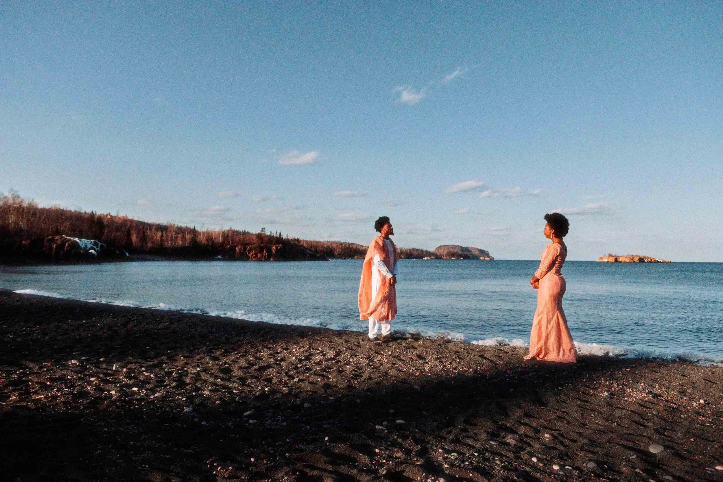 Two women standing on a dark sandy beach facing each other, with ocean water and overcast sky in the background.