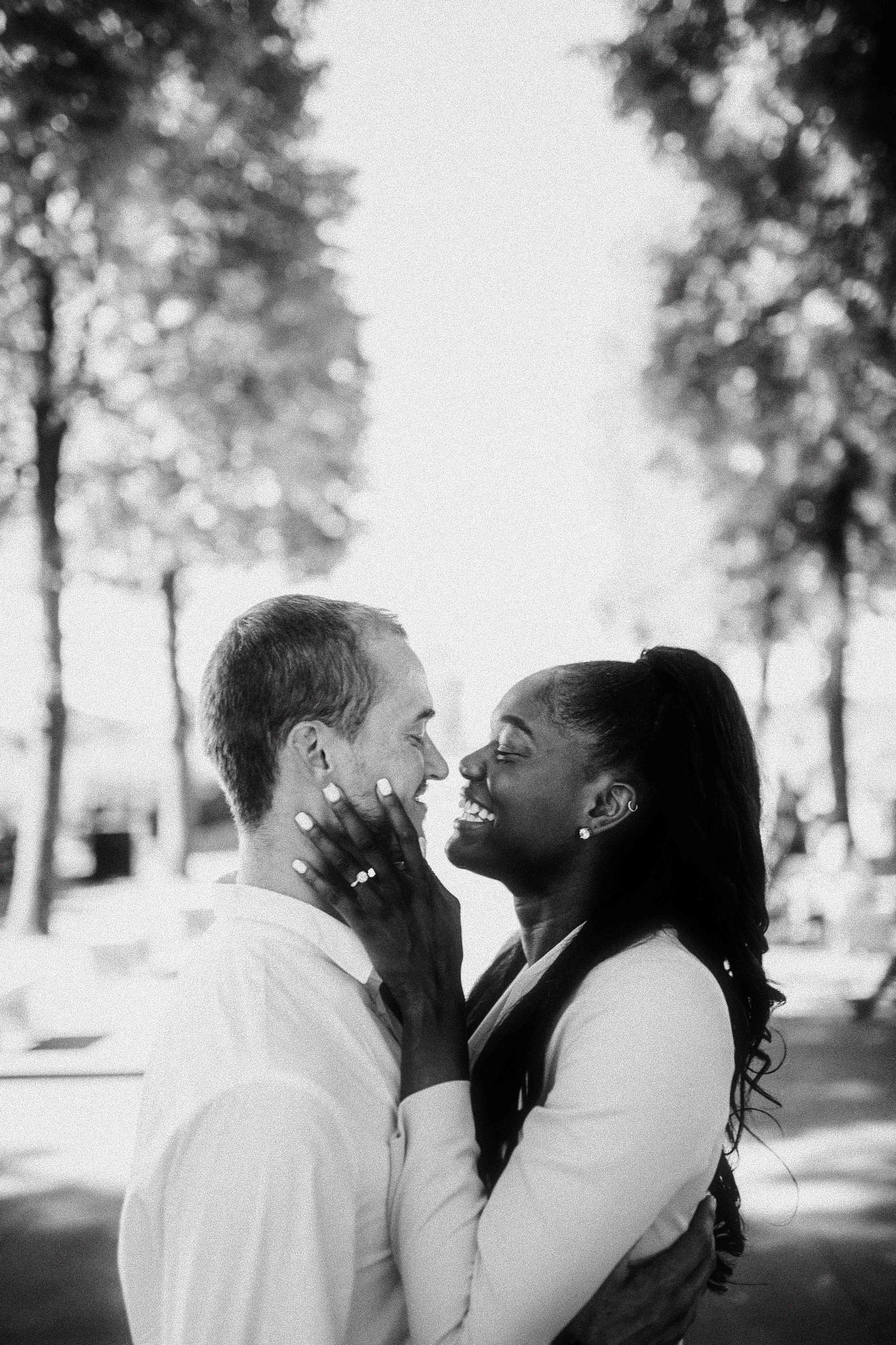 A black-and-white photo of a happy couple smiling and embracing outdoors, with trees in the background.