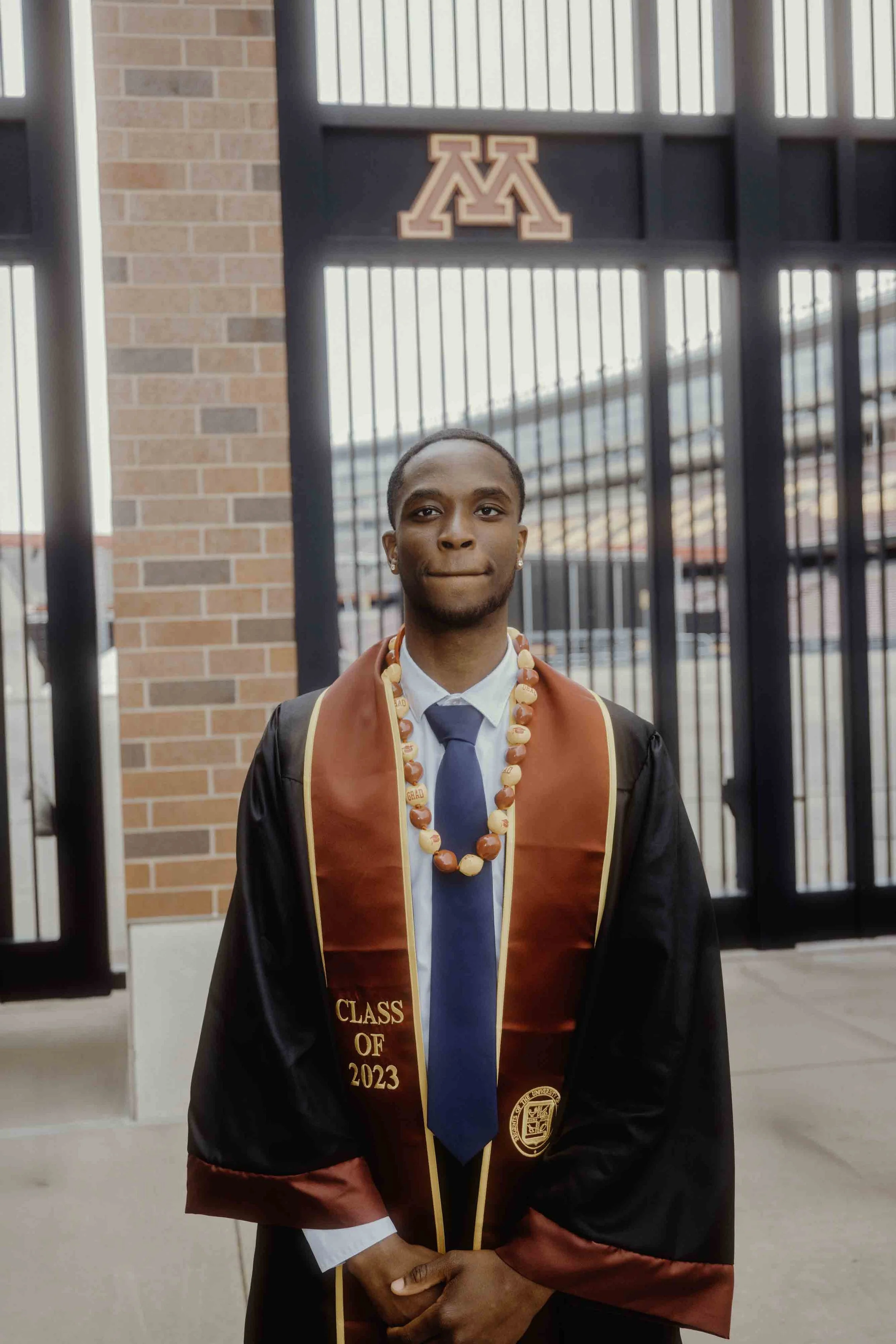 Graduate in black gown with red and gold stole, red and white beads, standing in front of a gate with a university logo.
