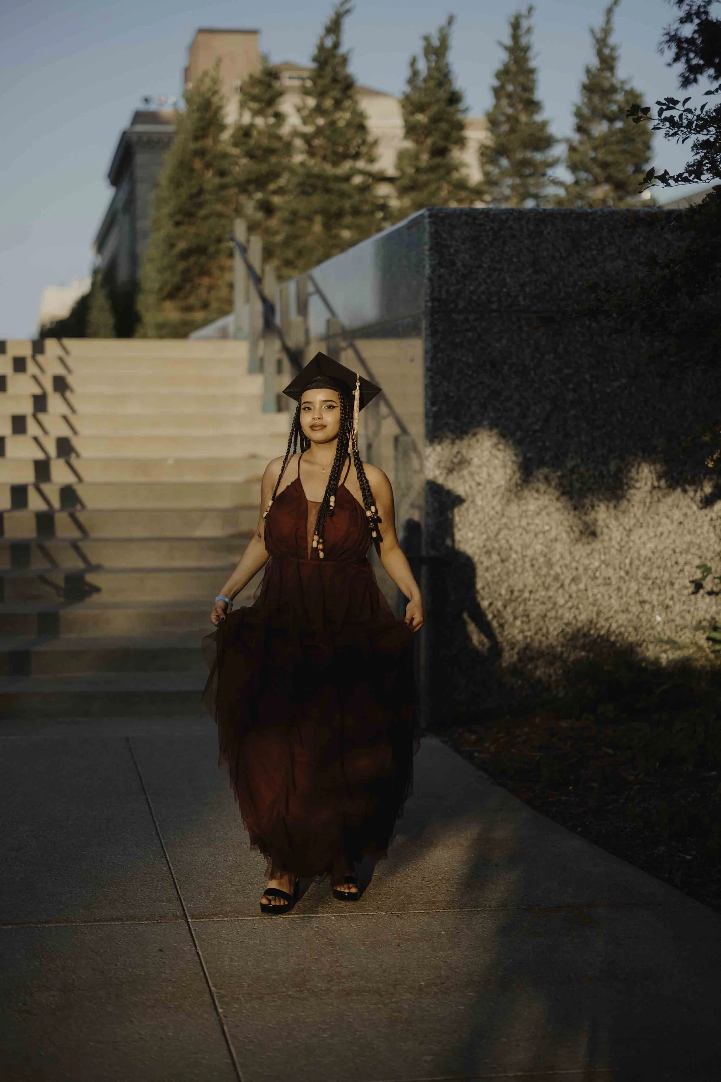 A young woman in a maroon dress and black graduation cap stands outdoors on a sidewalk, with steps and buildings in the background, during late afternoon or early evening sunlight.