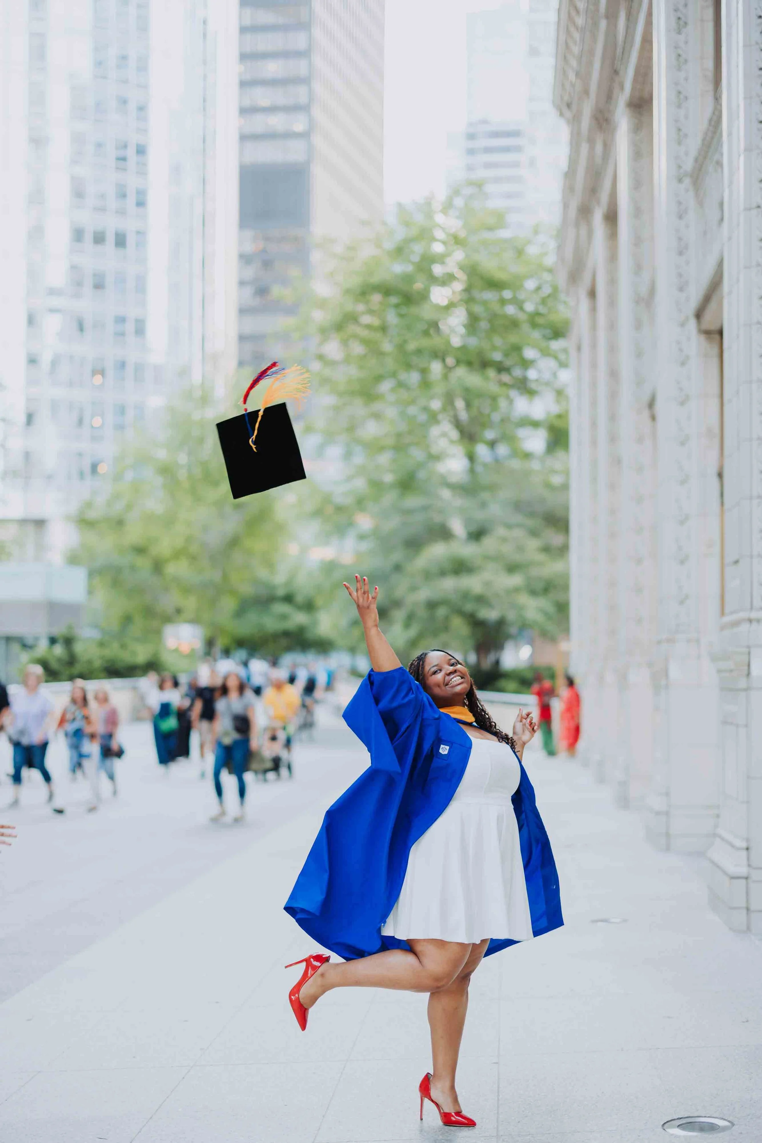A young woman in a white dress and blue graduation gown throws her cap in the air on a city street, smiling happily.
