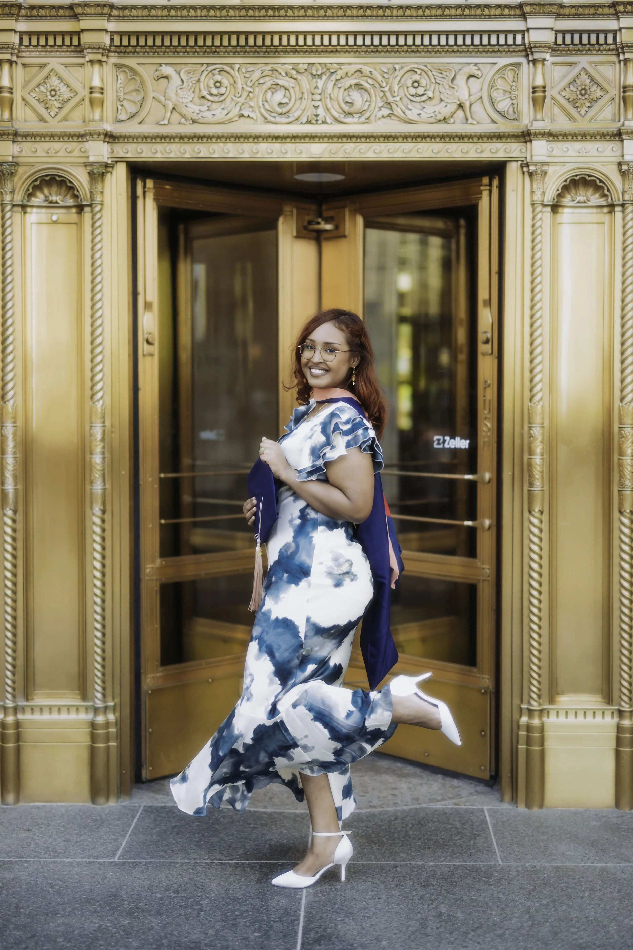 A woman wearing a blue and white patterned dress and white heels, smiling and holding a graduation cap, standing in front of ornate golden double doors.