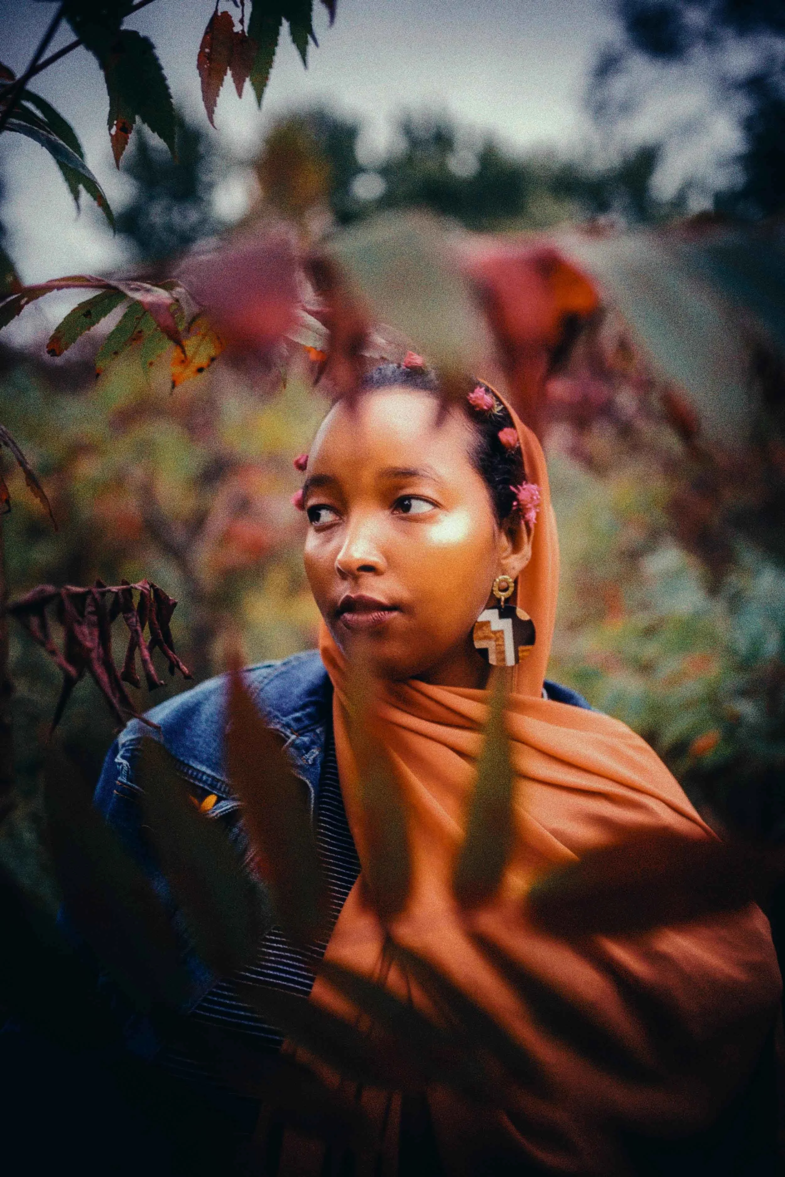 A woman with dark skin, short curly black hair decorated with pink flowers, wearing a large earring with geometric patterns, a peach headscarf, and a blue denim jacket, is surrounded by green and reddish foliage.