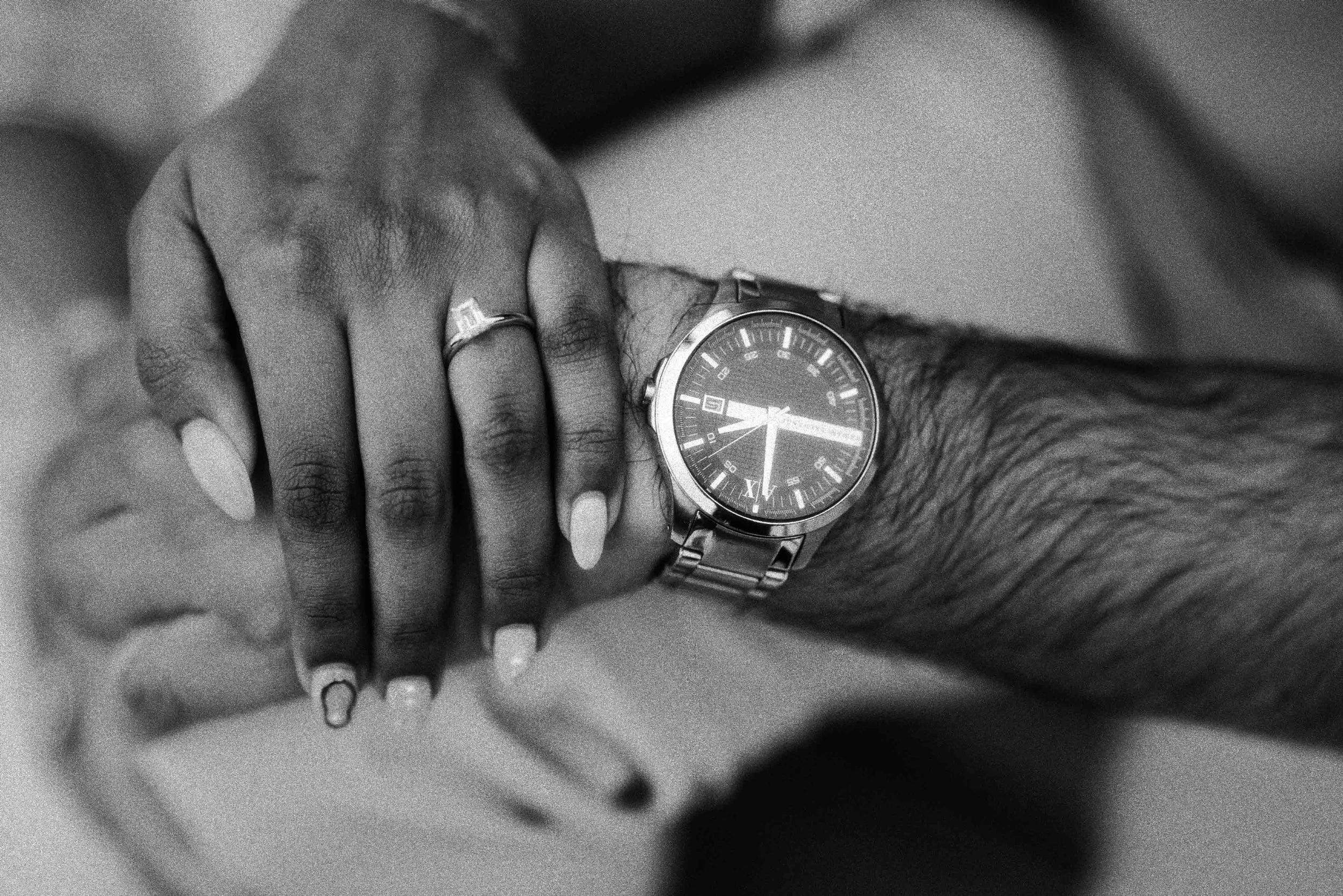 A close-up of a person's wrist wearing a silver wristwatch with a dark face and a metal band, along with another hand resting on top, showing rings and manicured nails.