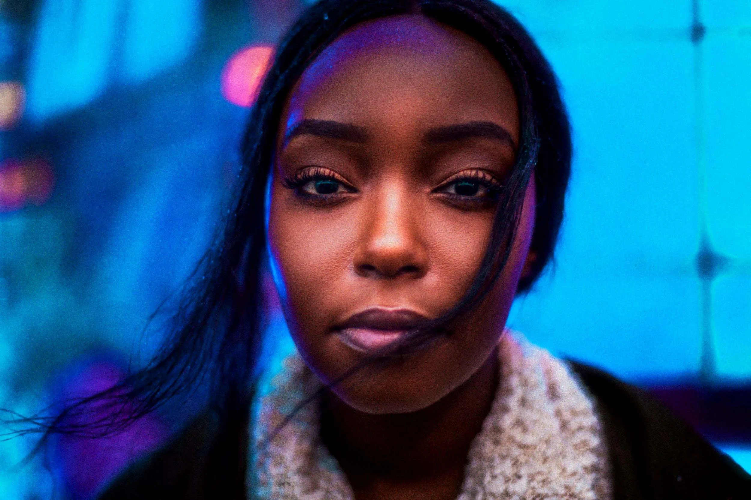 Close-up portrait of a young African American woman with straight black hair, wearing a beige textured sweater, with neon blue and pink lighting in the background.