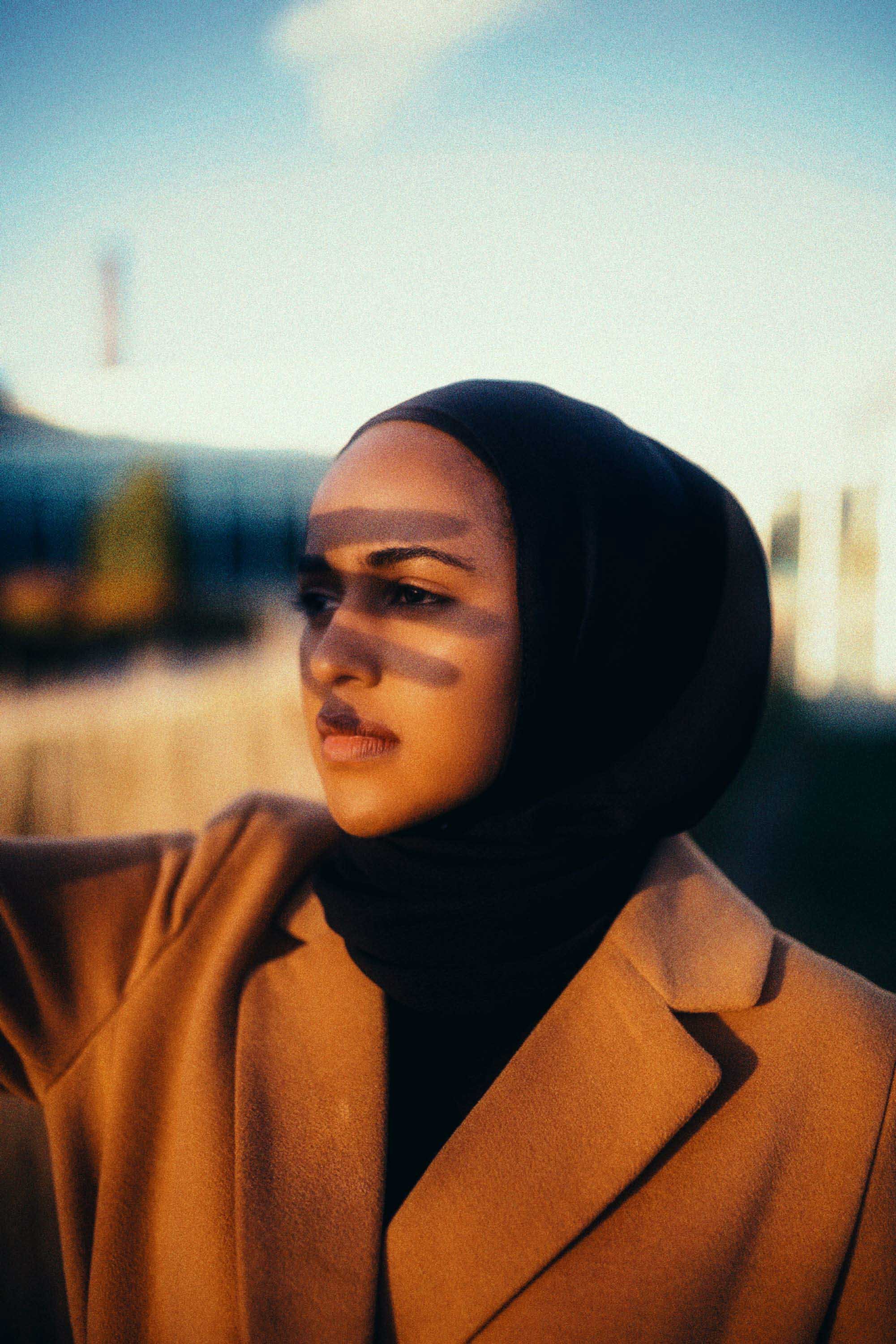 Close-up of a woman with a black headscarf and tan coat, looking into the distance with shadows on her face during sunset.