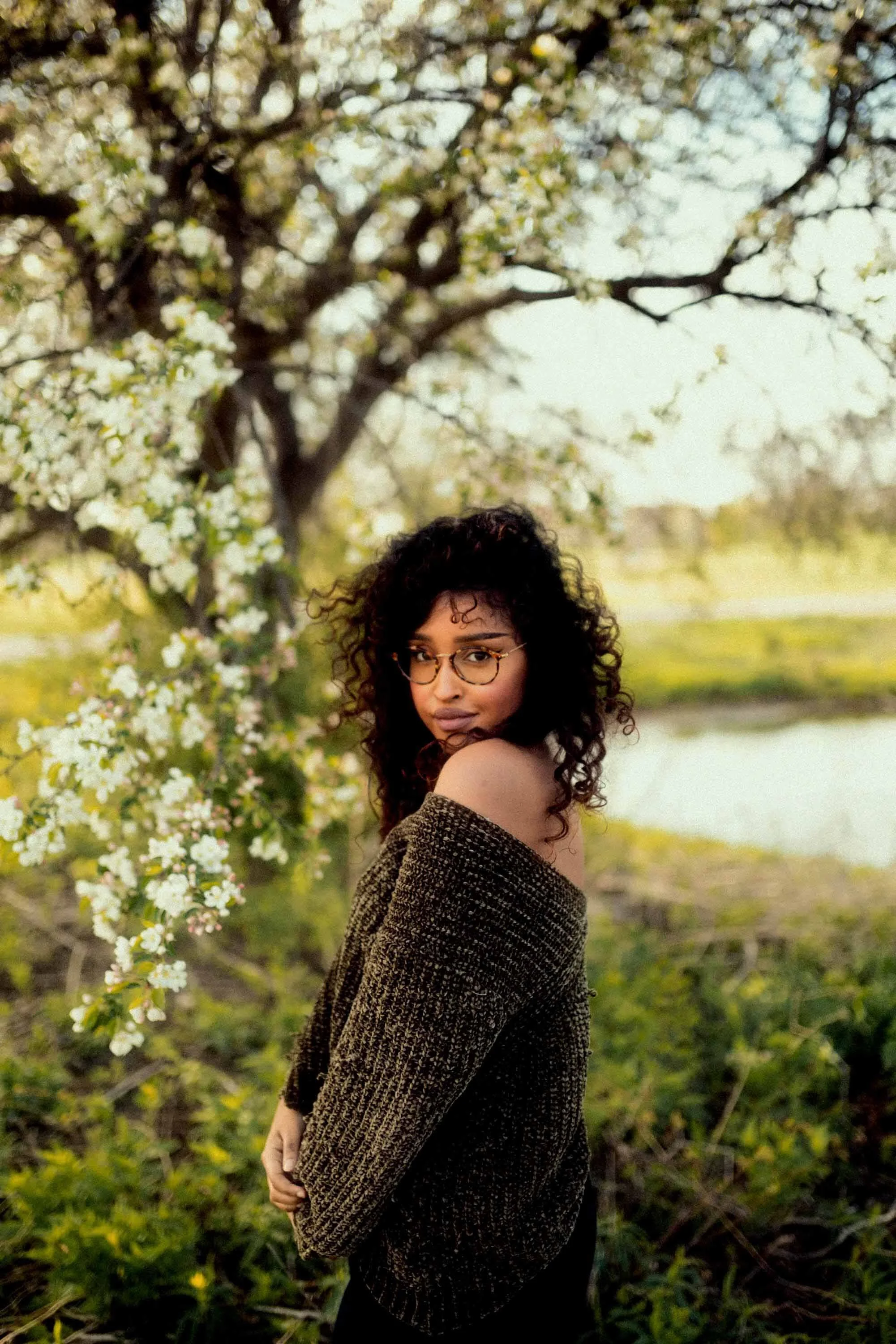 A woman with curly hair and glasses standing outdoors near a blossoming tree by a small body of water, wearing an off-the-shoulder sweater.