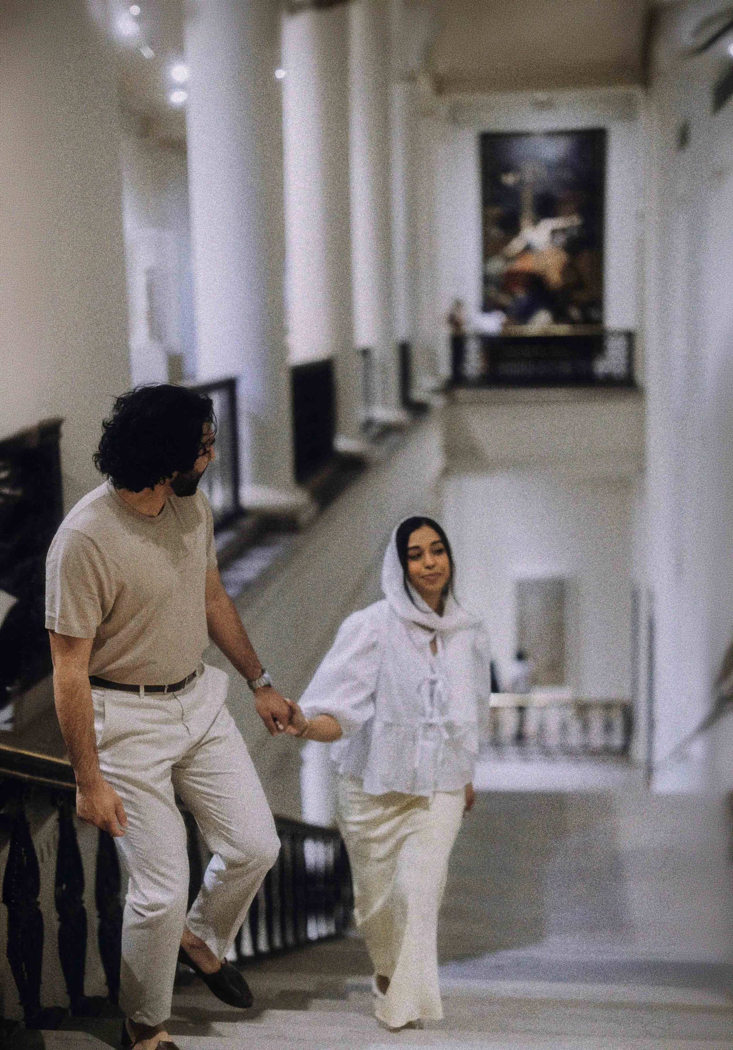 A man and woman holding hands and walking up a staircase in a well-lit, art-filled interior space.