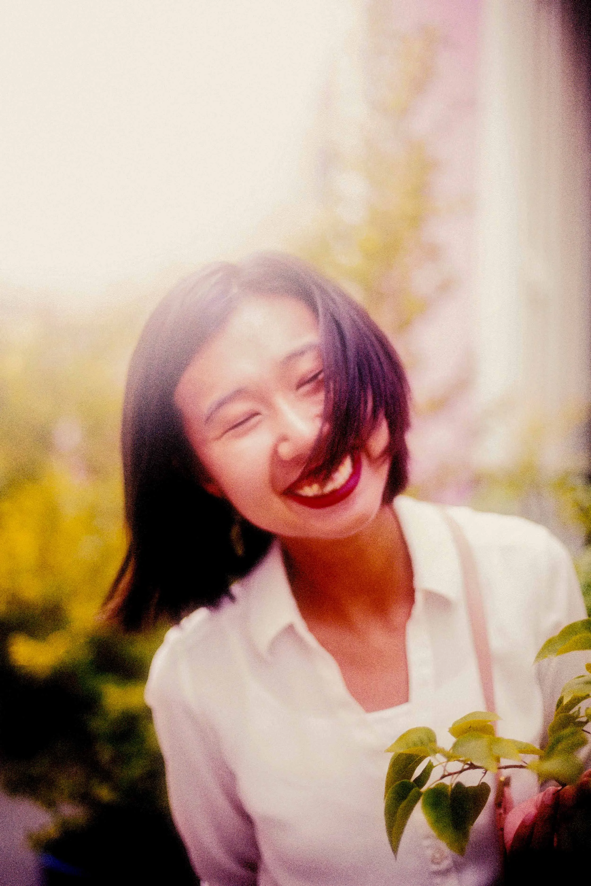 A young woman with short dark hair and a bright smile, wearing a white top, standing outdoors with blurred green foliage and sunlight in the background.