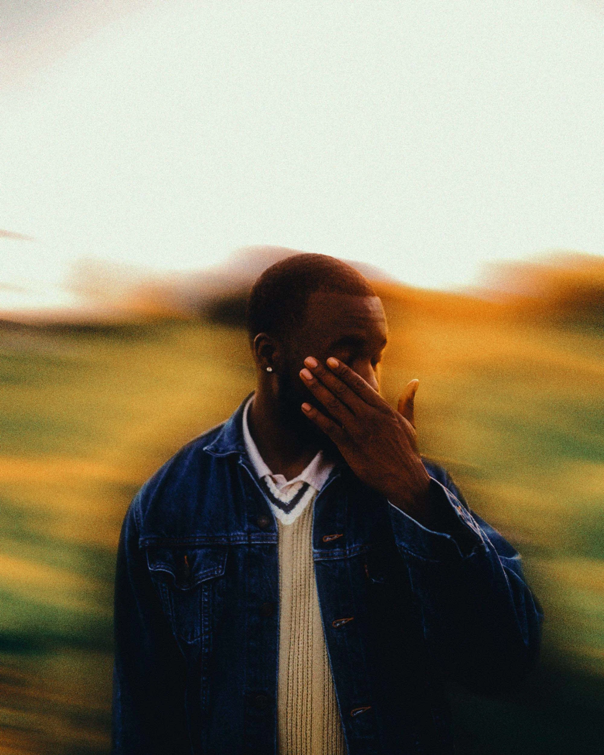 A person with short hair wearing a denim jacket and sweater, standing outdoors with a blurred background of green and orange foliage, touching their face.