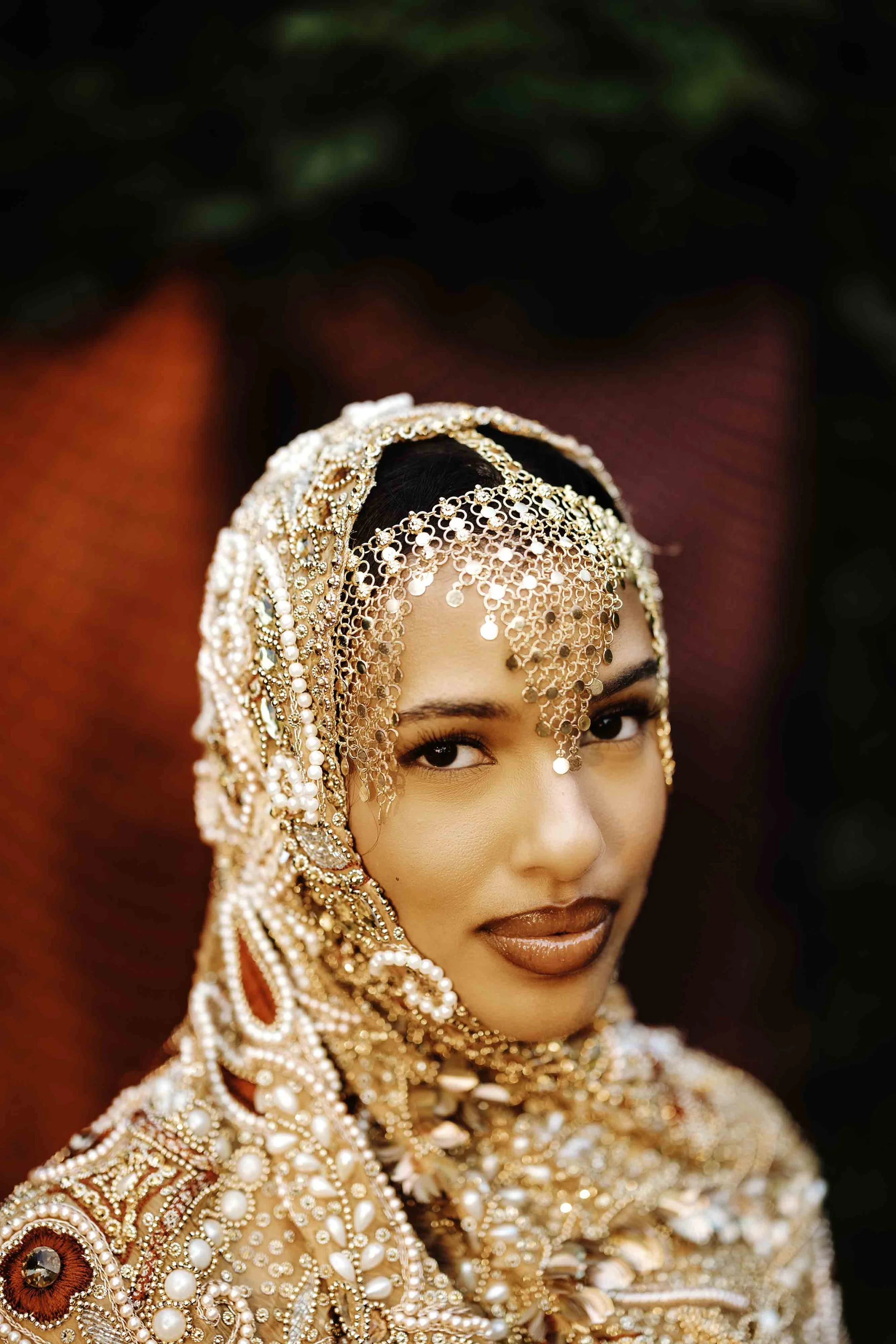 A woman dressed in ornate gold and pearl bridal attire with intricate jewelry, including a bejeweled headpiece, with a red and purple backdrop.