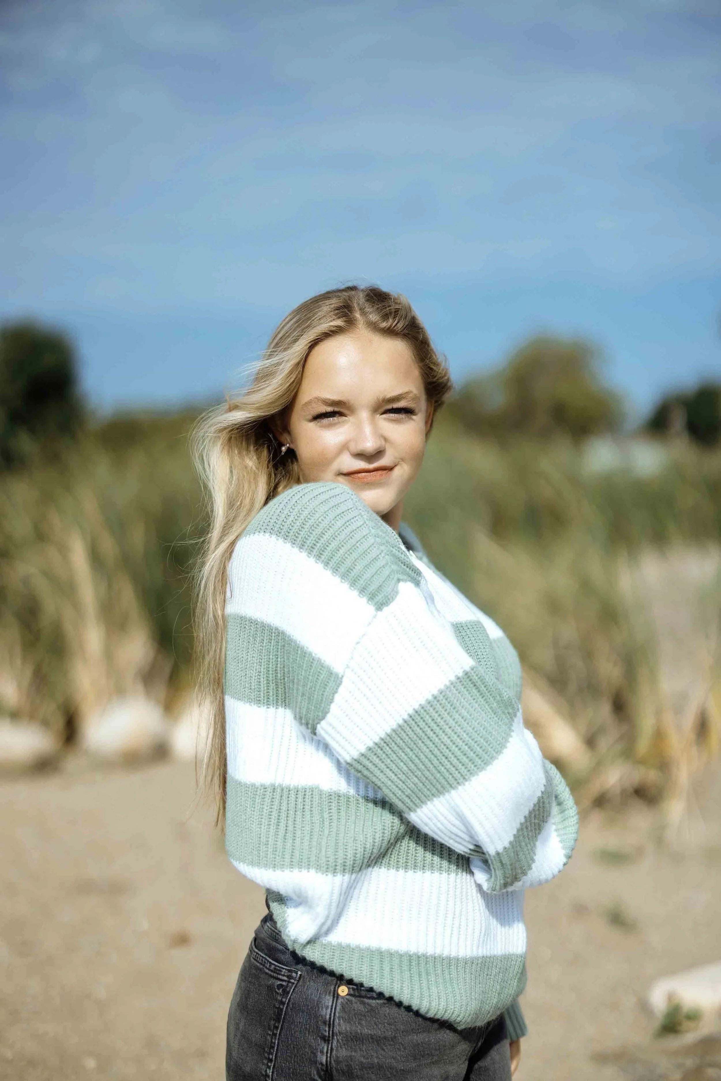 A young woman with long blonde hair standing outdoors on a sunny day, wearing a green and white striped sweater and dark jeans, with a blurred natural background behind her.