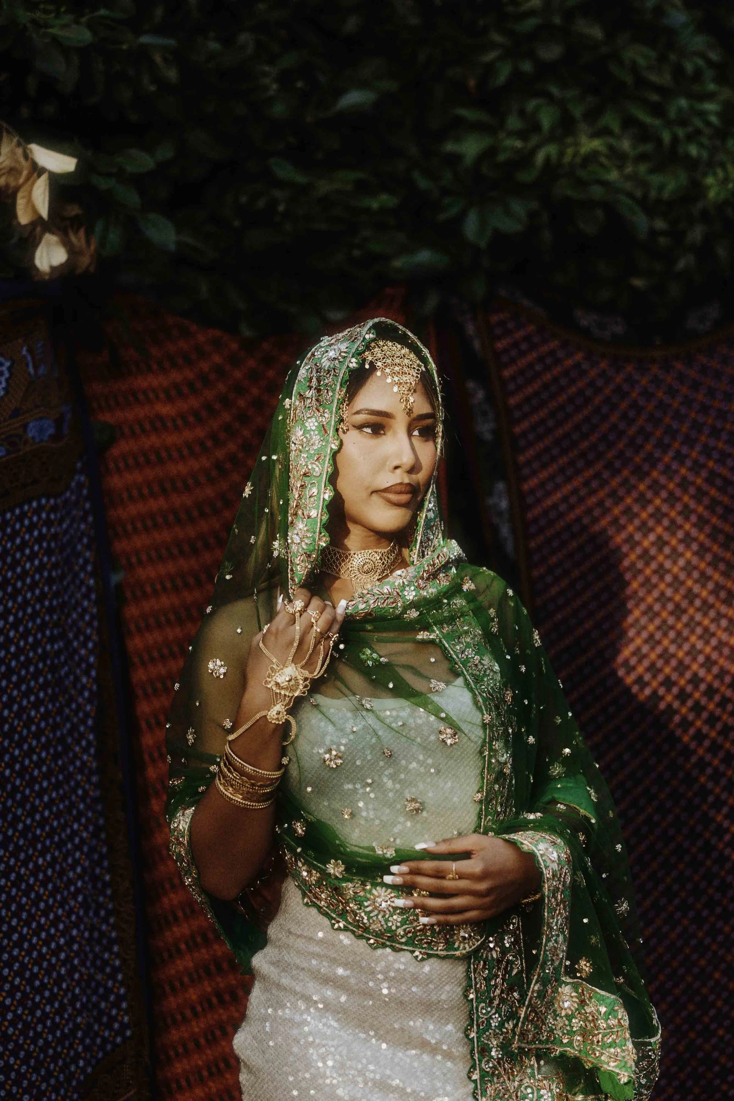 A woman in traditional Indian attire with green and gold embroidery, wearing jewelry, standing against a backdrop of colorful fabrics.