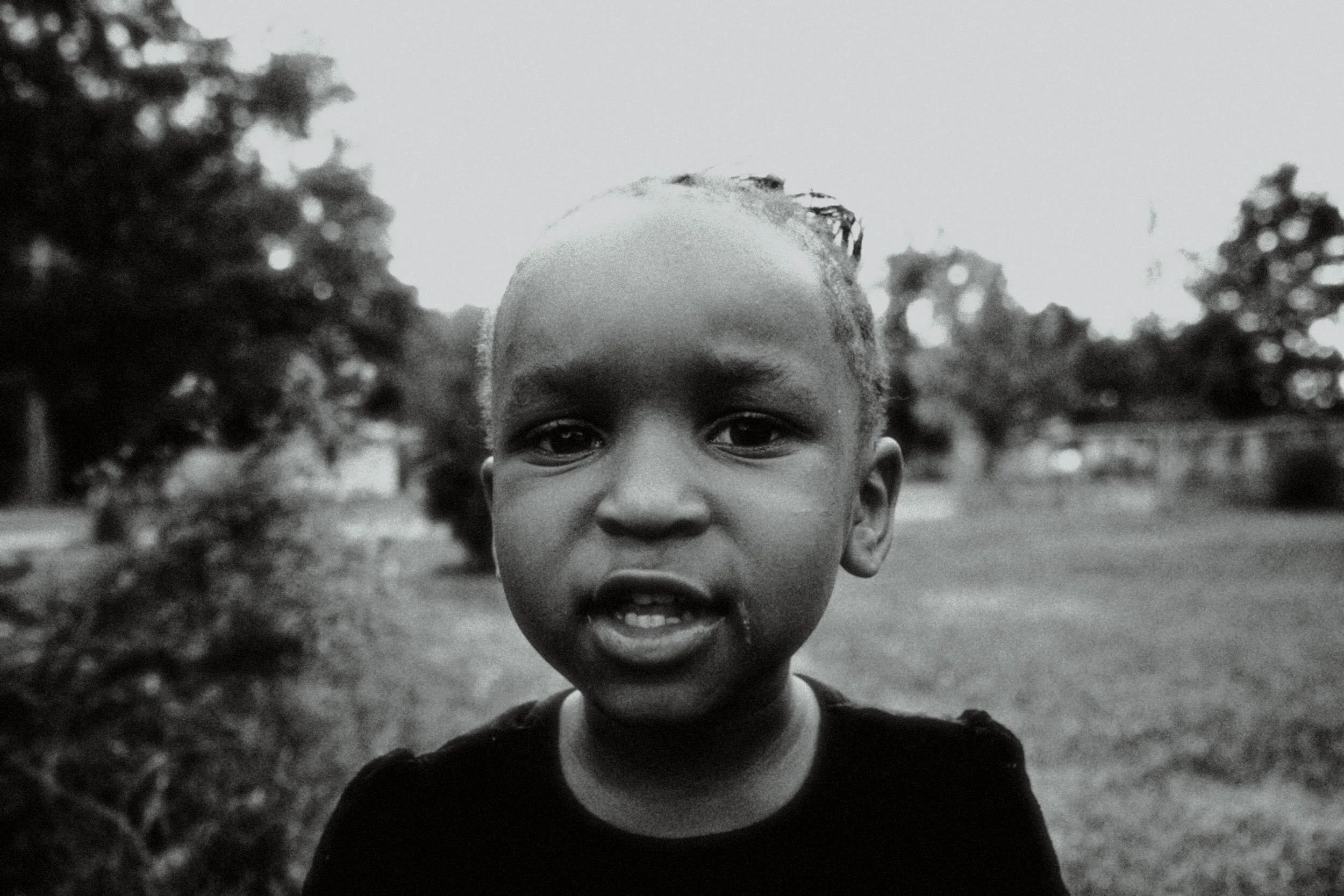 A young boy outdoors in a park or garden area, with trees and grass in the background, looking directly at the camera in black and white.