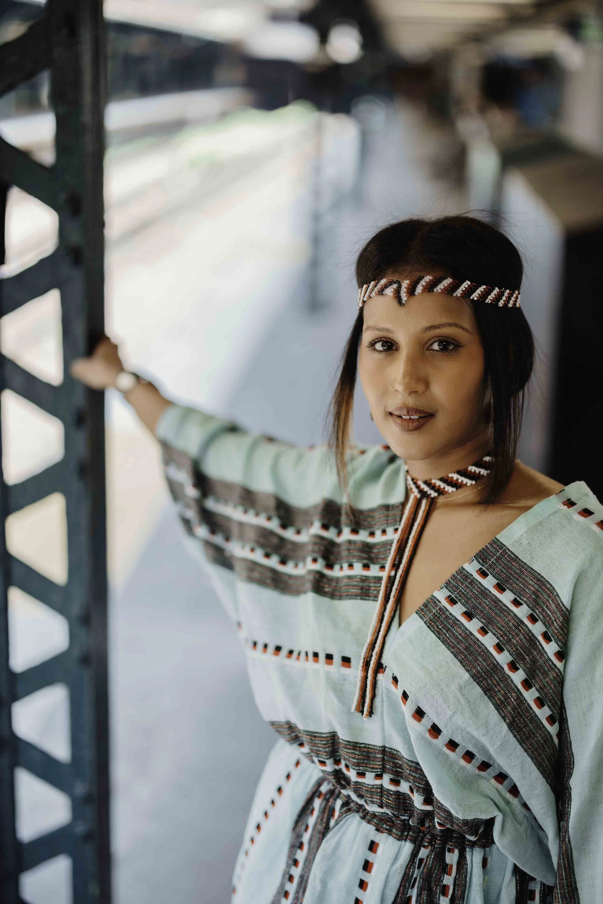 A young woman dressed in traditional clothing with a patterned headband, standing next to a metal structure, looking at the camera with a slight smile.