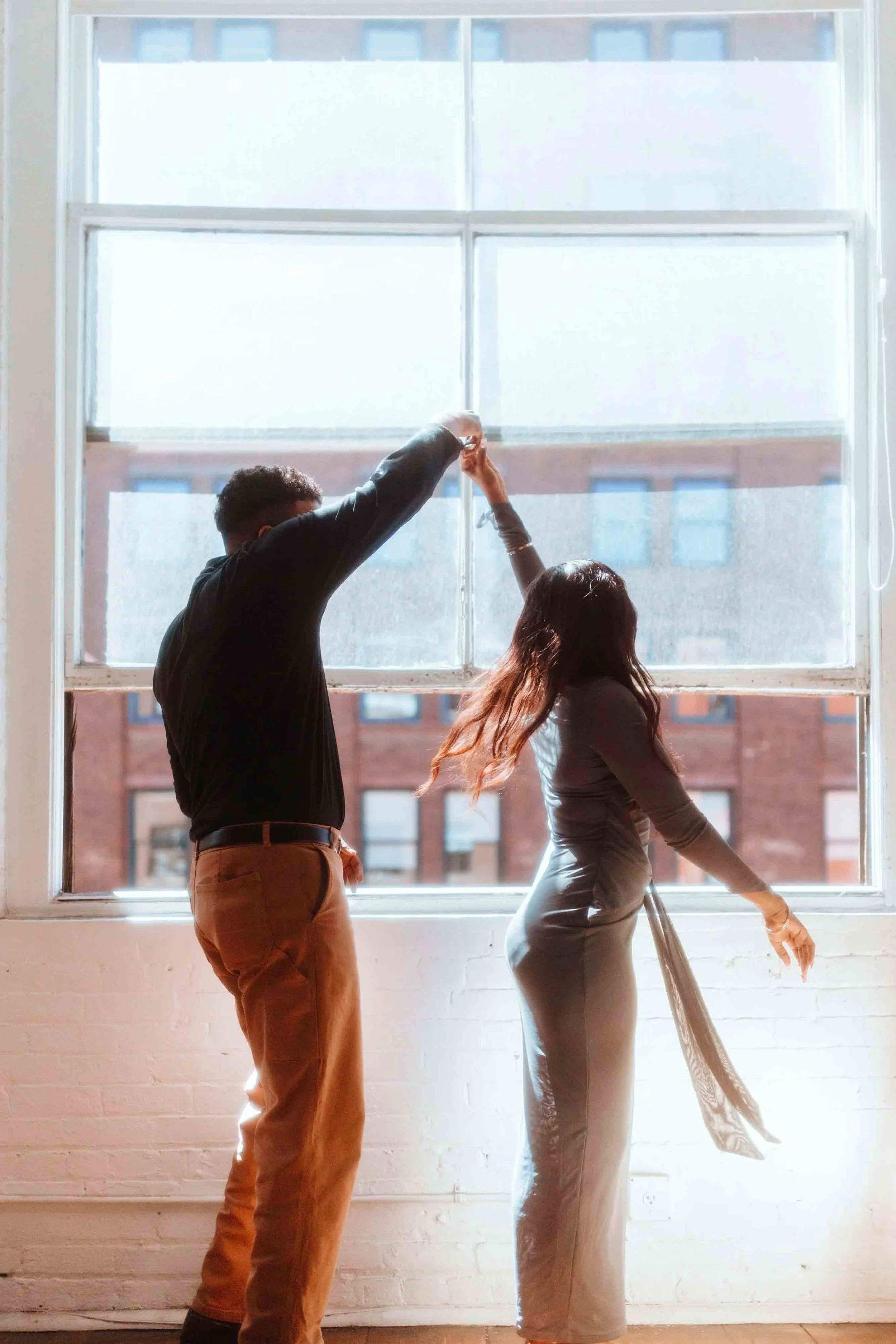 A man and woman dancing in front of a large window with sunlight shining through, with a brick building visible outside.