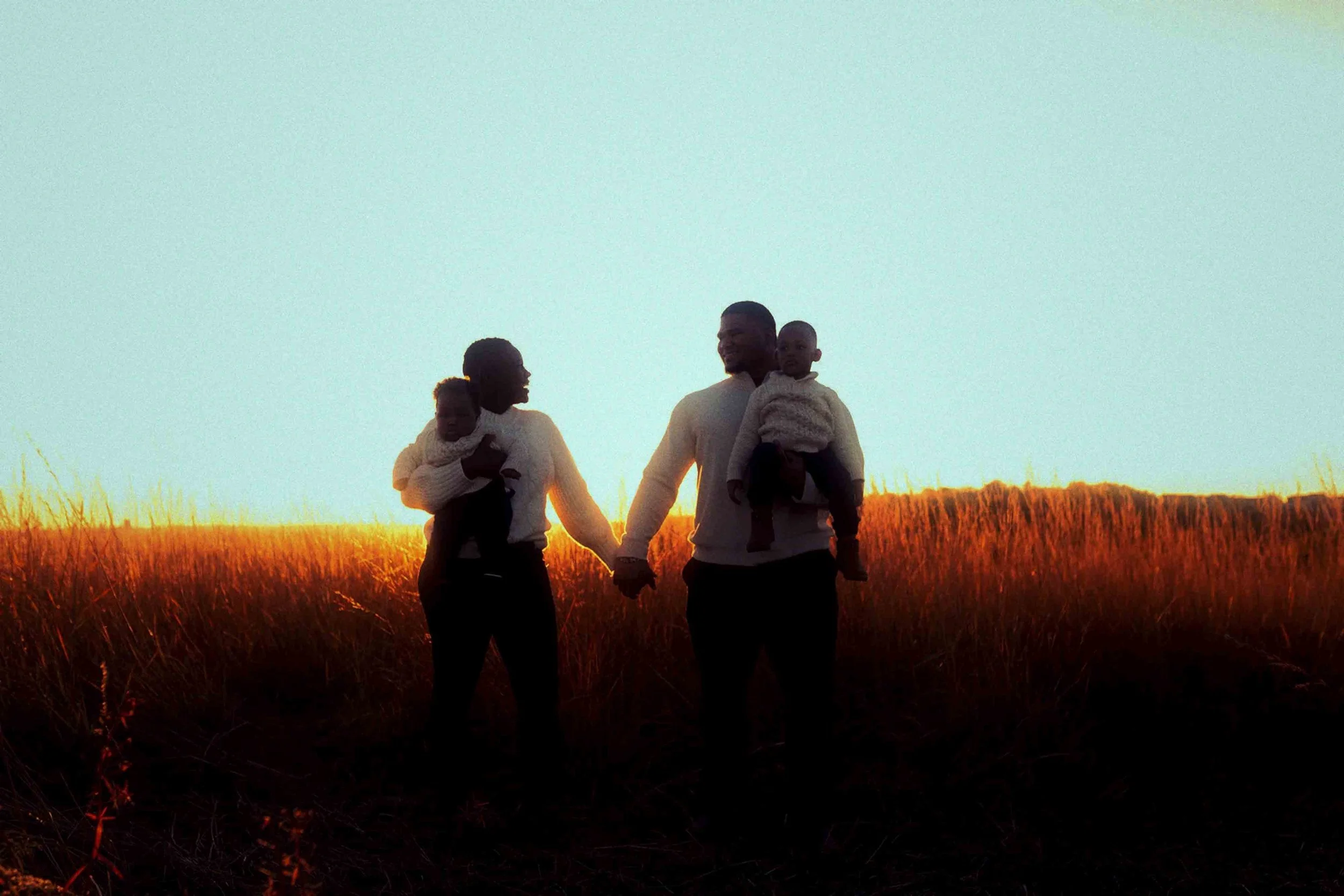 A family of two adults and two children holding hands and walking through a grassy field at sunset.