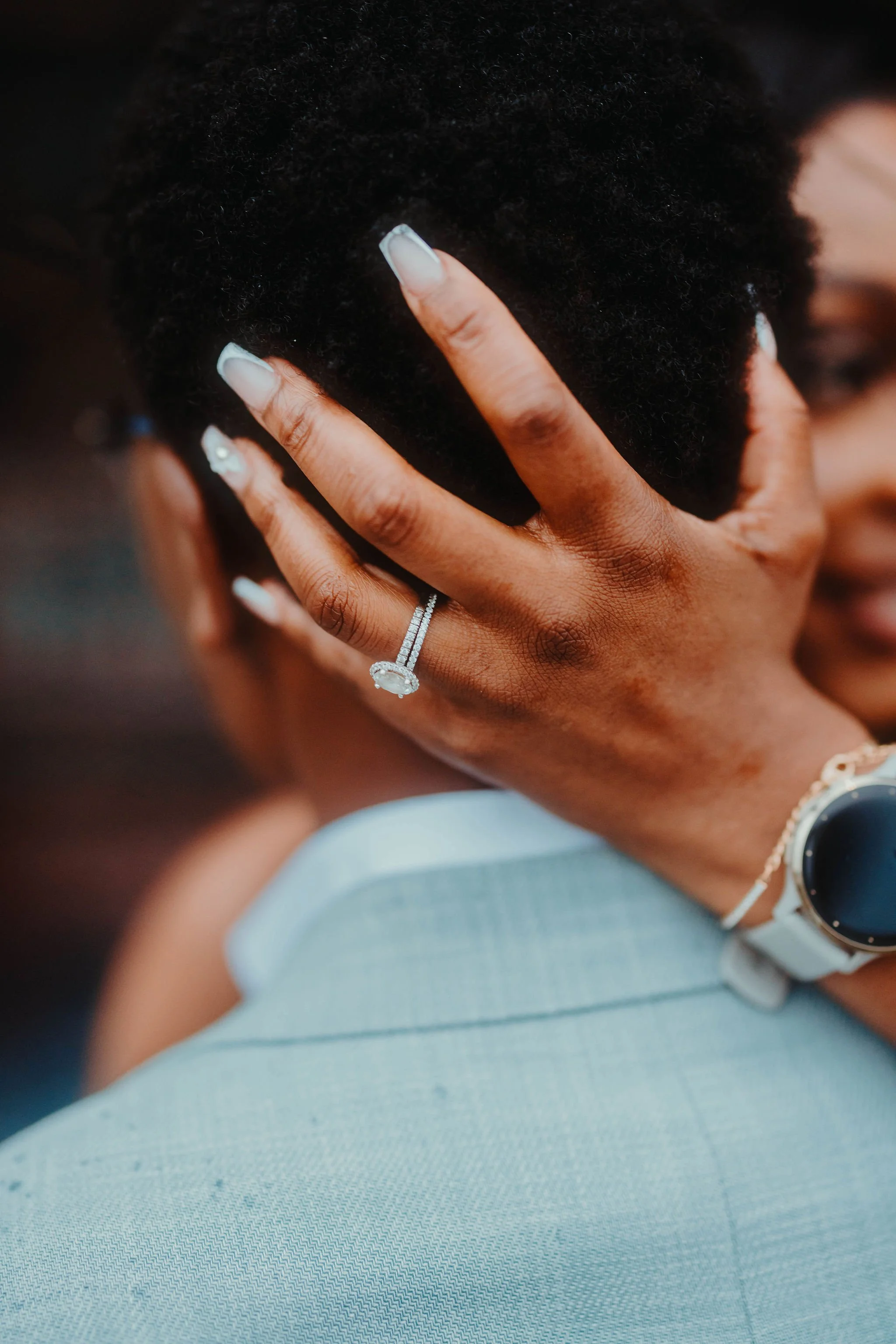 Close-up of a woman’s face being held gently by a hand with an engagement ring, showing her white manicure, wearing a white watch and bracelet, with dark curly hair, and wearing a light-colored blazer.