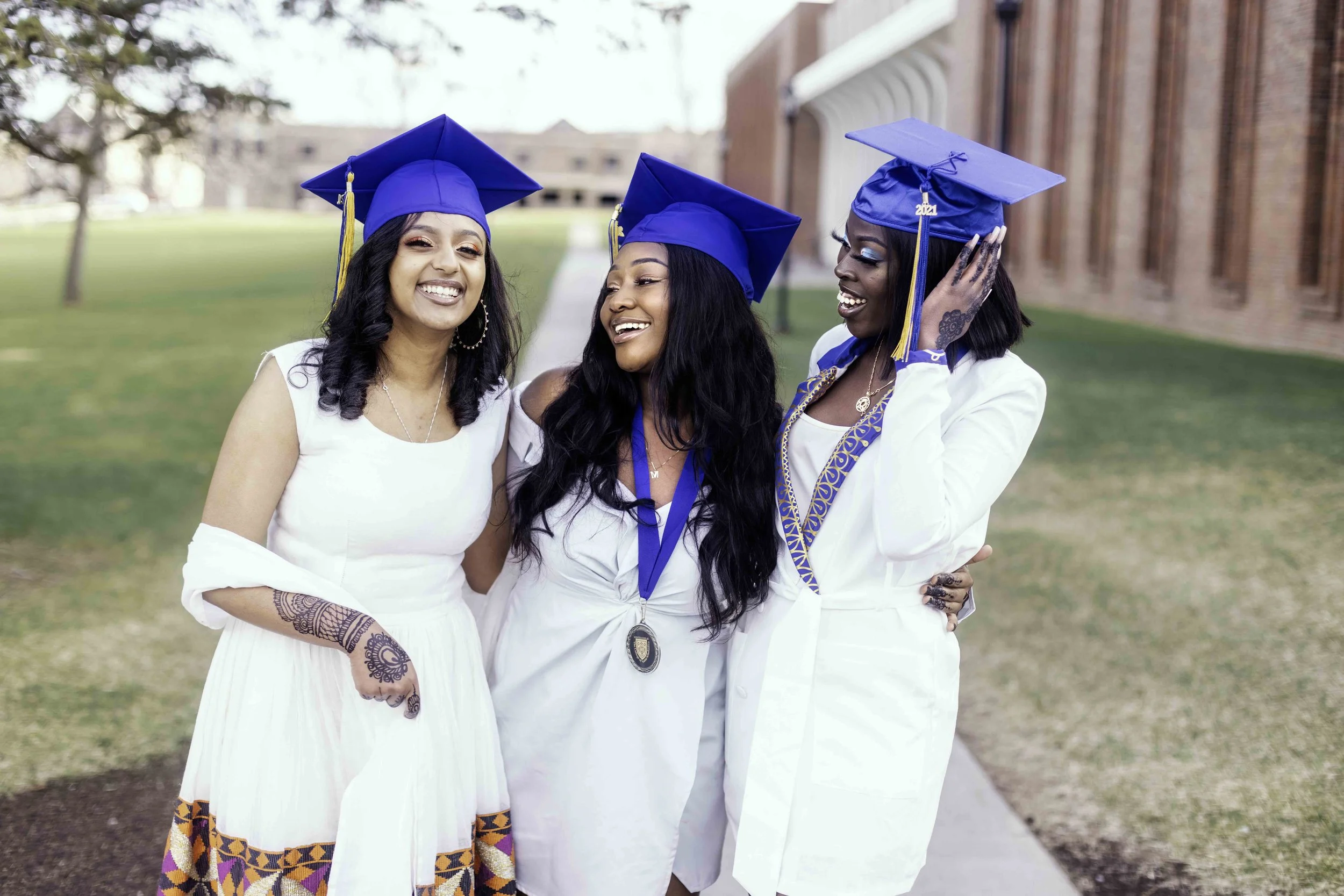 Three women in graduation caps and gowns romanticize joyfully outside on a campus, smiling and posing together.