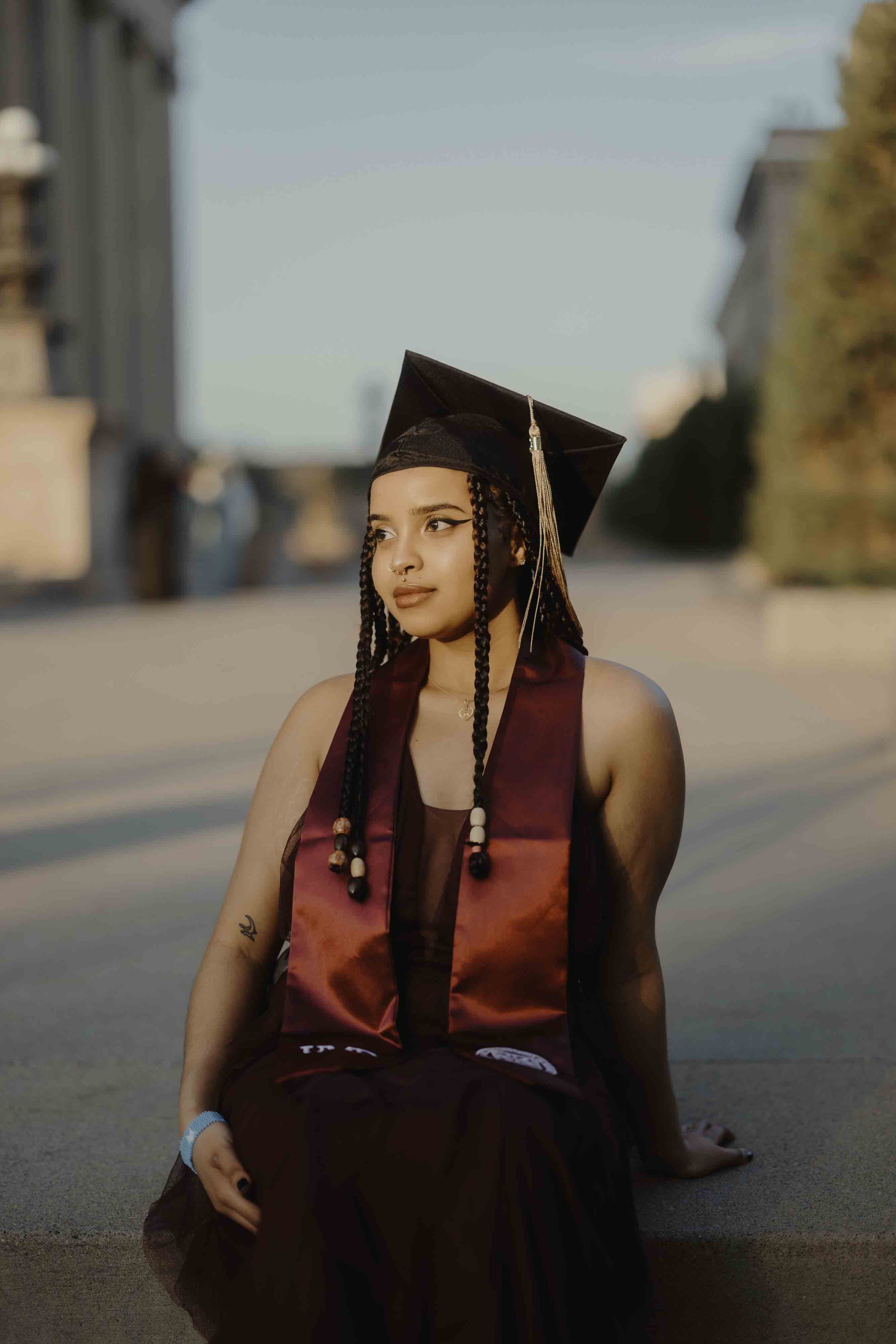 Young woman in graduation cap and gown sitting outdoors on a sunny day, with a city street and buildings in the background.