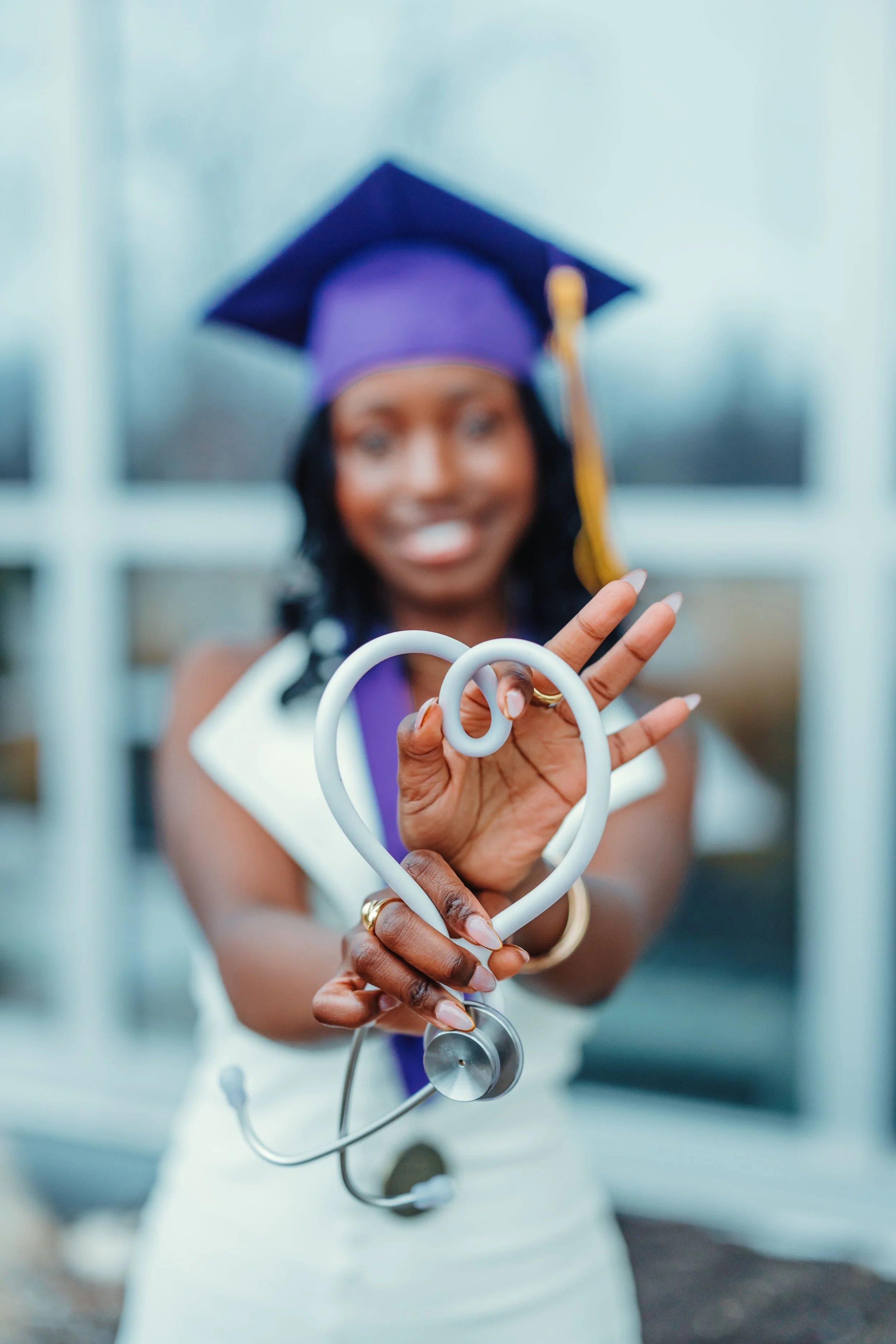 Woman wearing a graduation cap and gown holding a stethoscope shaped like a heart, smiling at the camera.