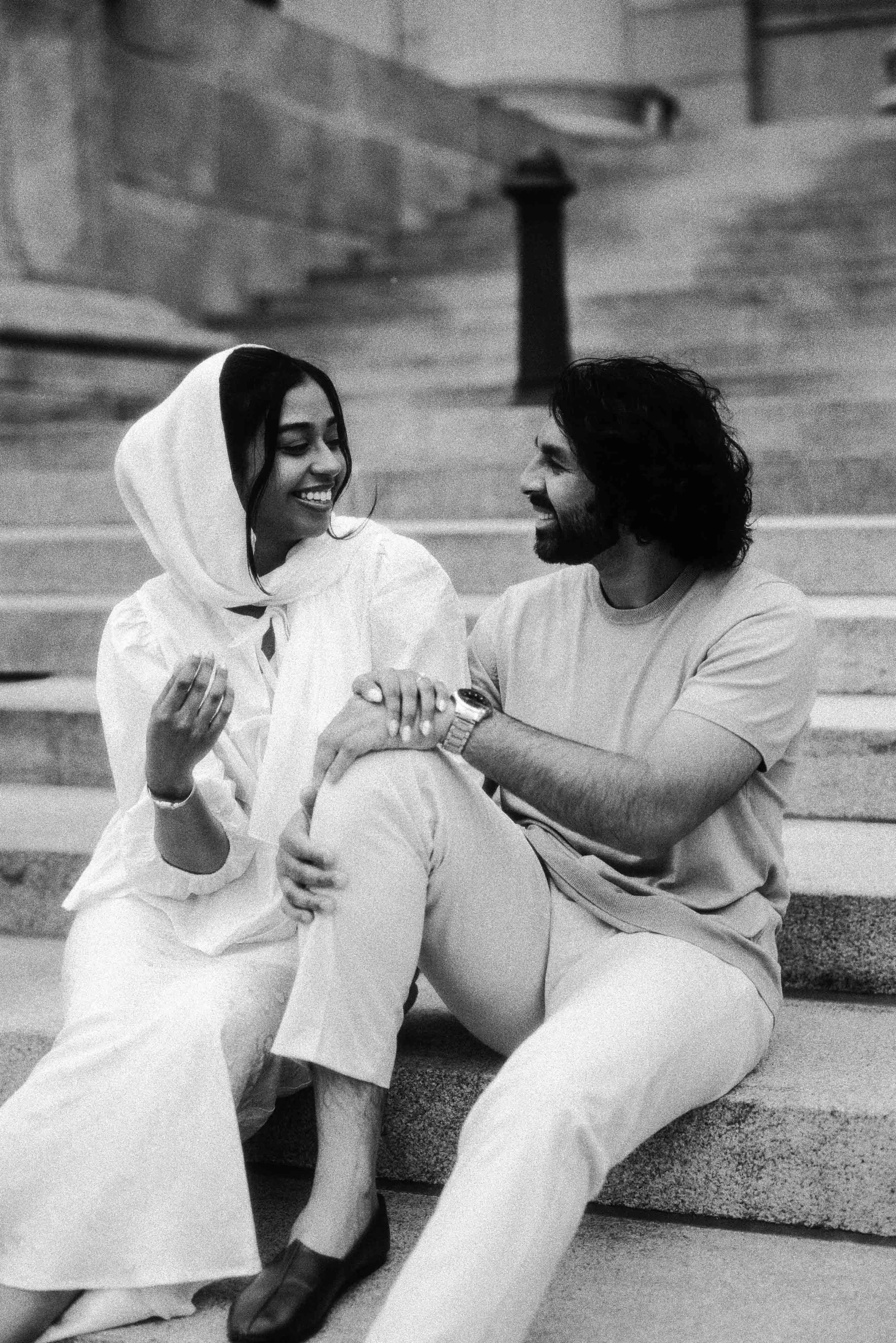 A woman and a man sitting on stairs, smiling and looking at each other. The woman is wearing a hoodie and the man has long hair and a beard.