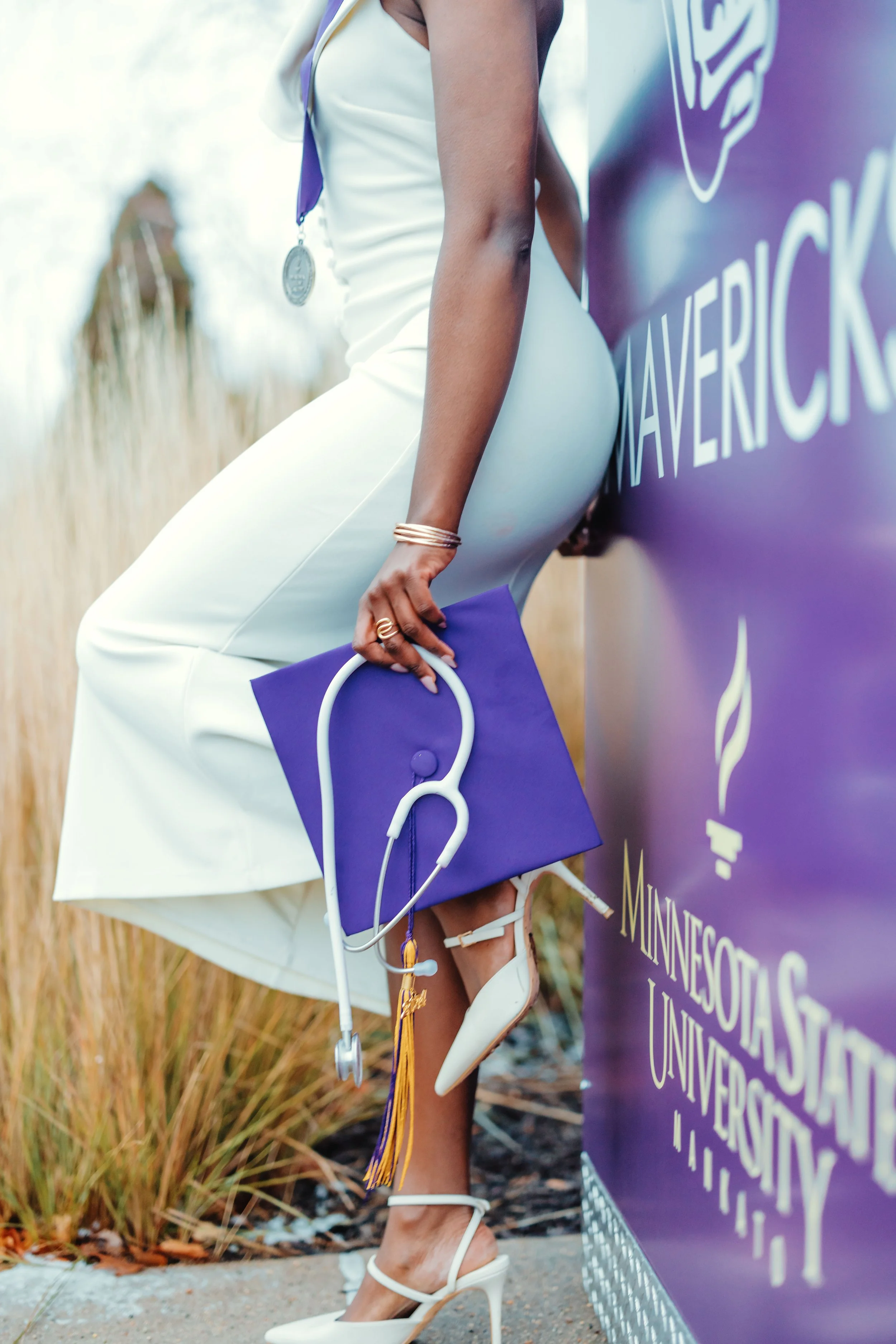 Close-up of a woman in white high heels and white dress holding a purple clutch with a stethoscope hanging from it, standing next to a purple sign for Minnesota State University.