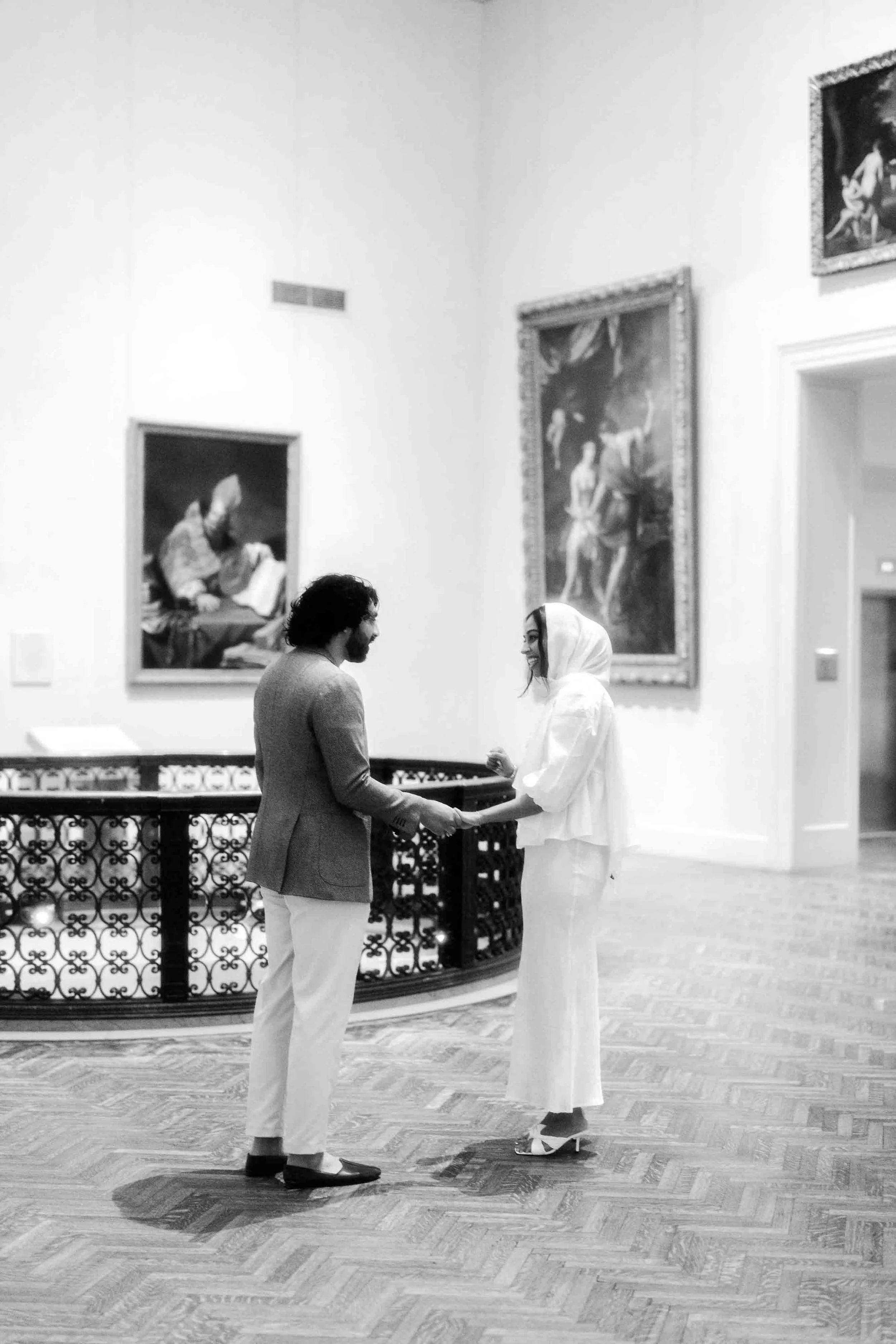 A black and white photo of a man and woman holding hands and smiling at each other in an art gallery. The woman is wearing a white outfit and a headscarf, and the man is dressed in a blazer and light-colored pants. They are standing on a wooden floor