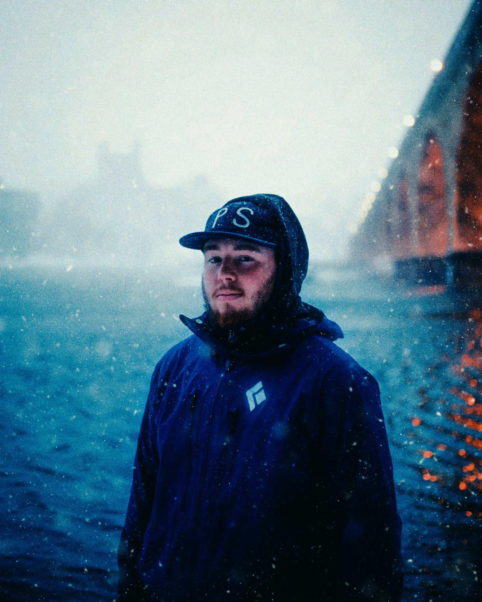 A young man with a beard standing in a snowy, foggy setting with a body of water and a blurred bridge in the background, wearing a black cap and jacket.