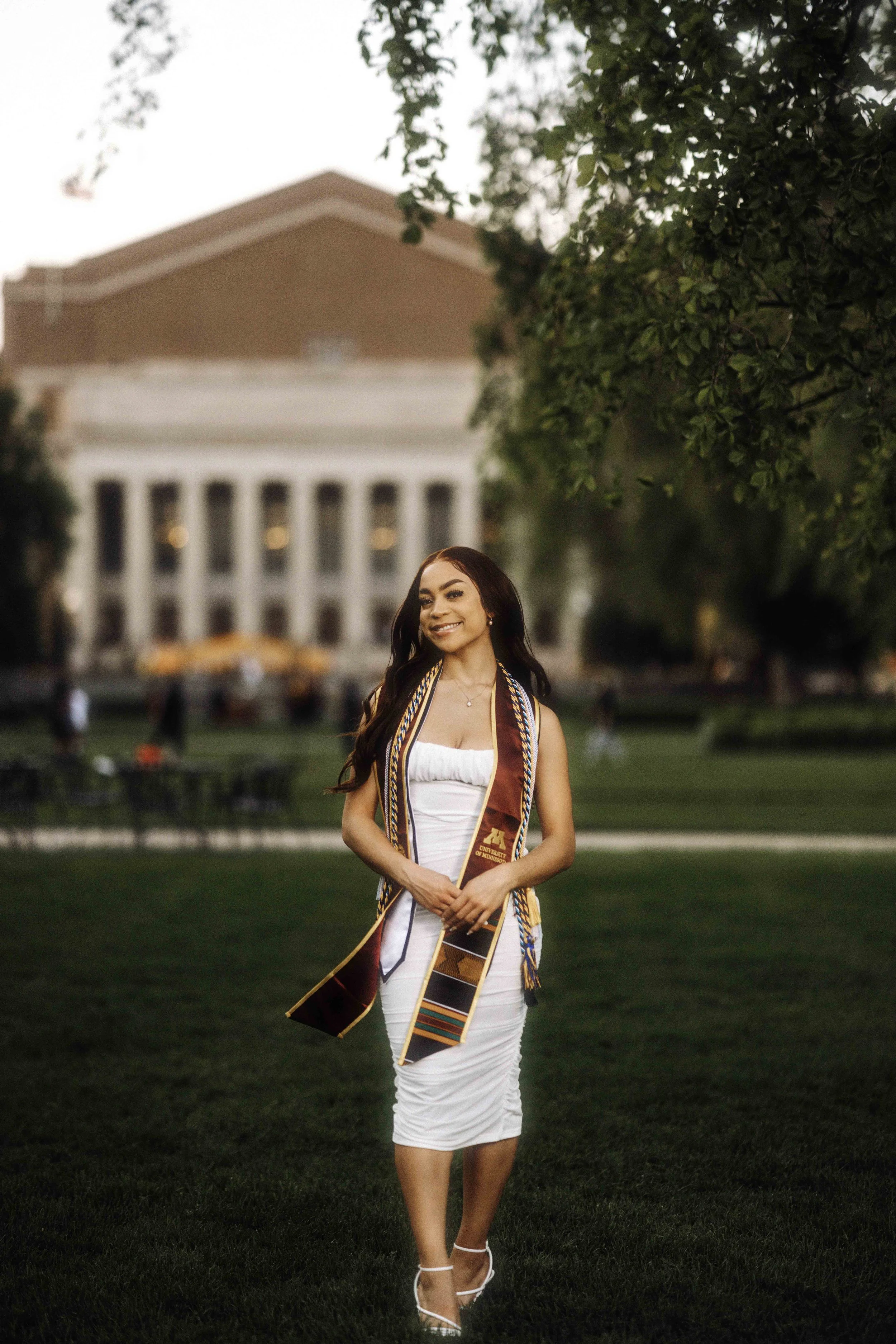 Young woman in white dress and graduation stole standing on grass in front of college campus.