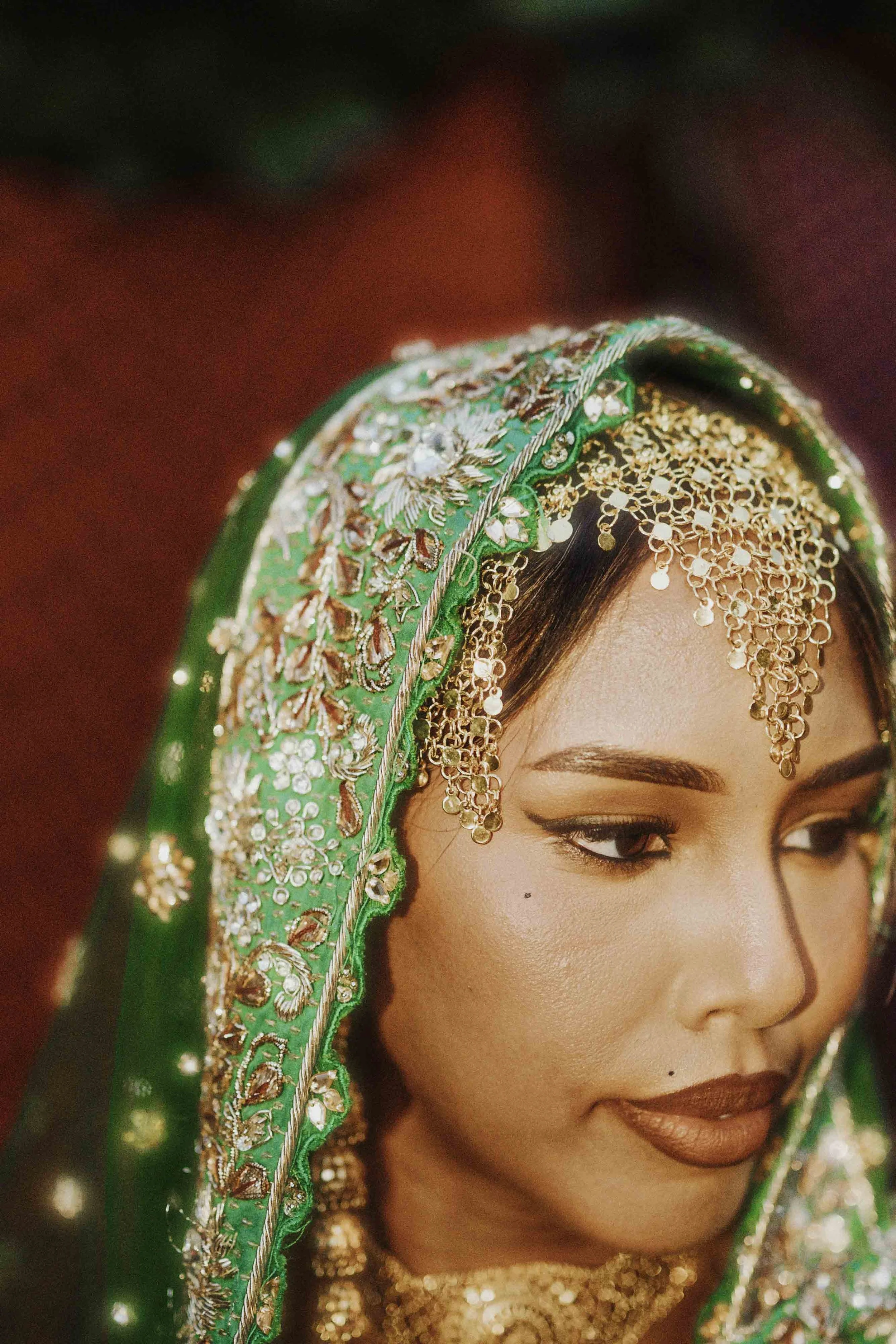 Close-up of a woman in traditional Indian bridal attire wearing elaborate gold jewelry, including a headpiece and earrings, with detailed embroidery on her green veil.