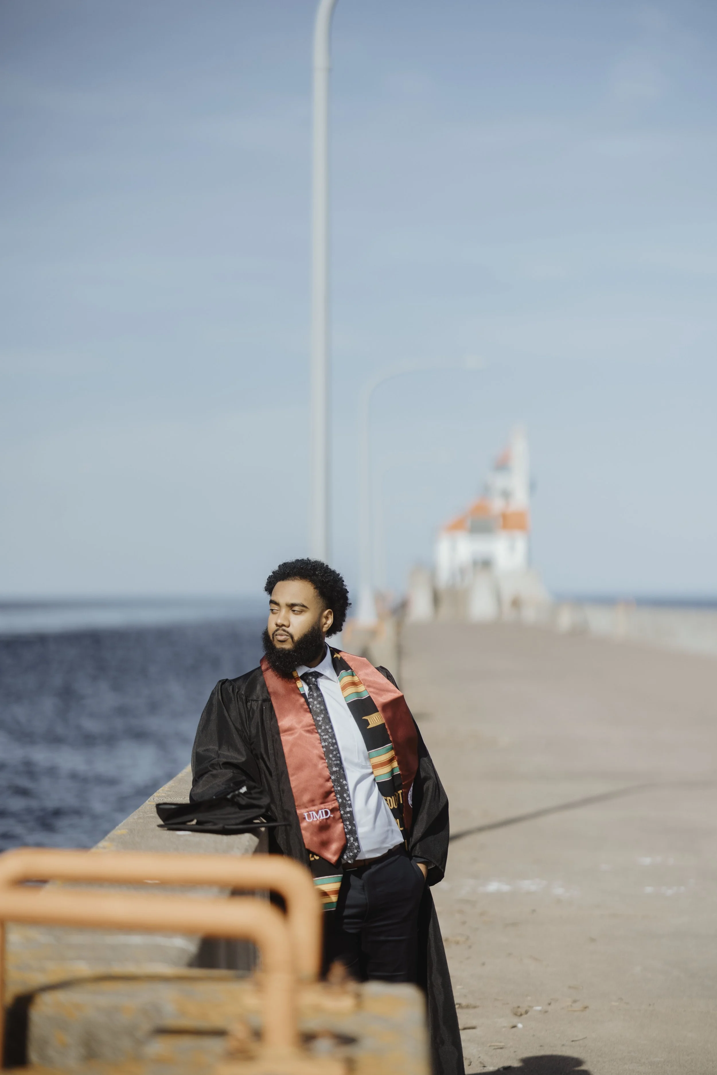 A man wearing a graduation gown and stole standing on a pier, with a lighthouse in the background.