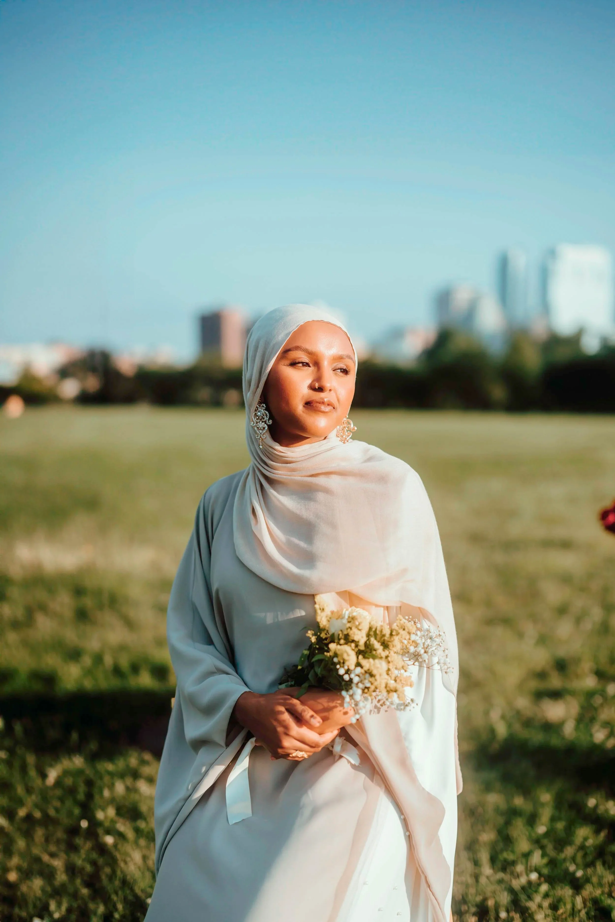 Woman wearing a cream-colored hijab and dress holding a small bouquet of white flowers in a green field with city buildings in the background during sunset.
