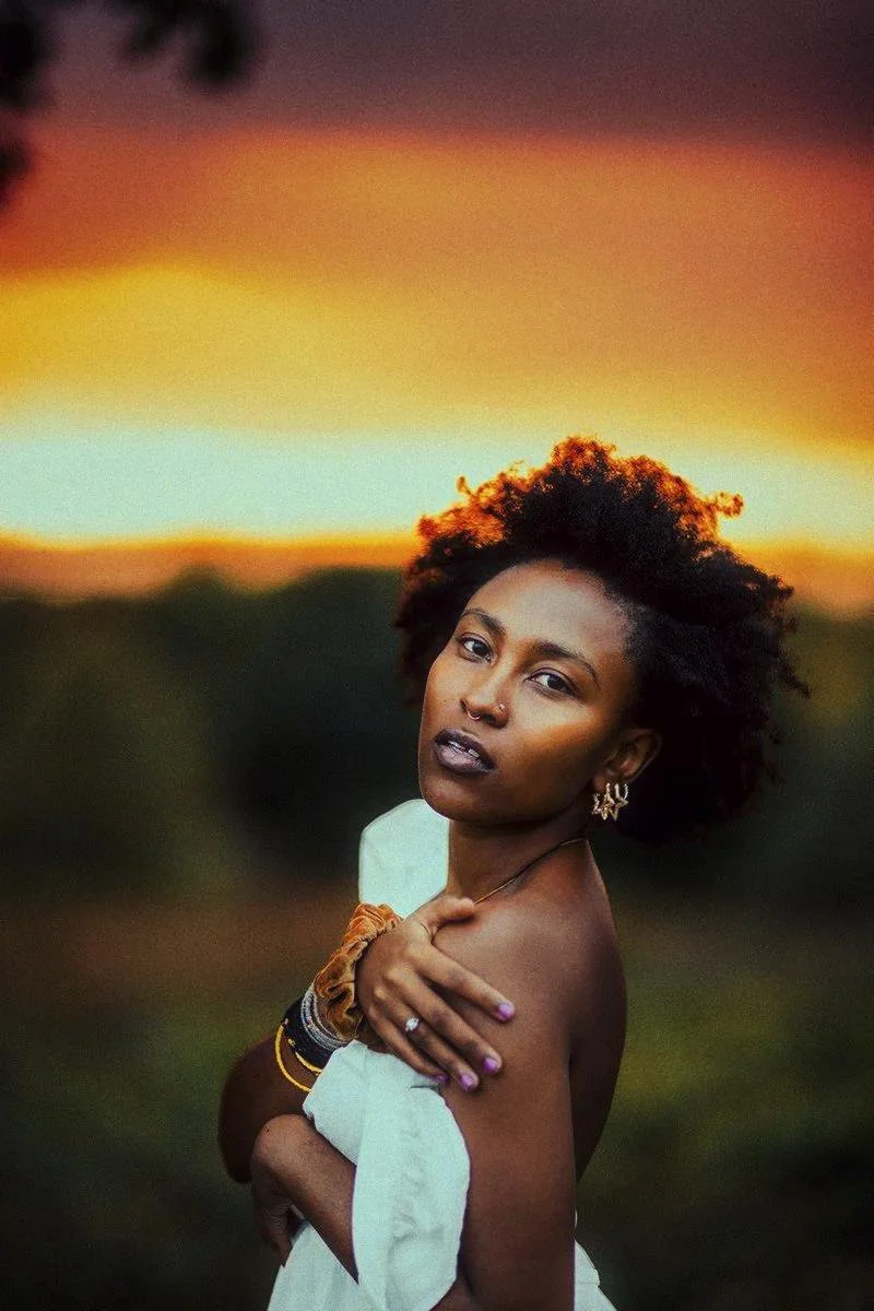 A woman with dark skin and natural curly hair posing outdoors at sunset. She has a septum piercing, multiple bracelets, and earrings, and is wearing a white off-the-shoulder garment.