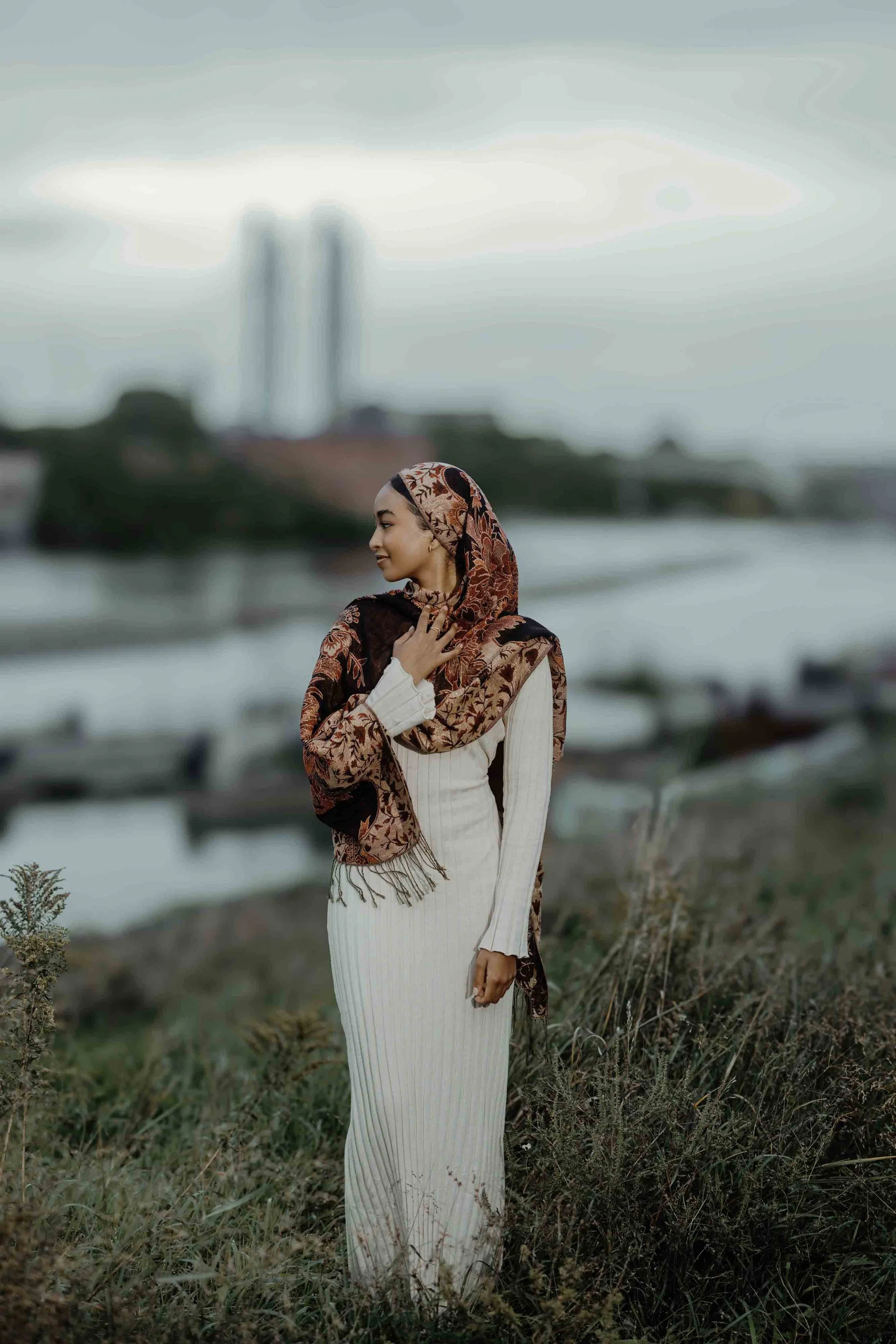 A woman wearing a white dress and a red and black patterned hijab standing in a field near water with boats, with a blurred cityscape in the background.