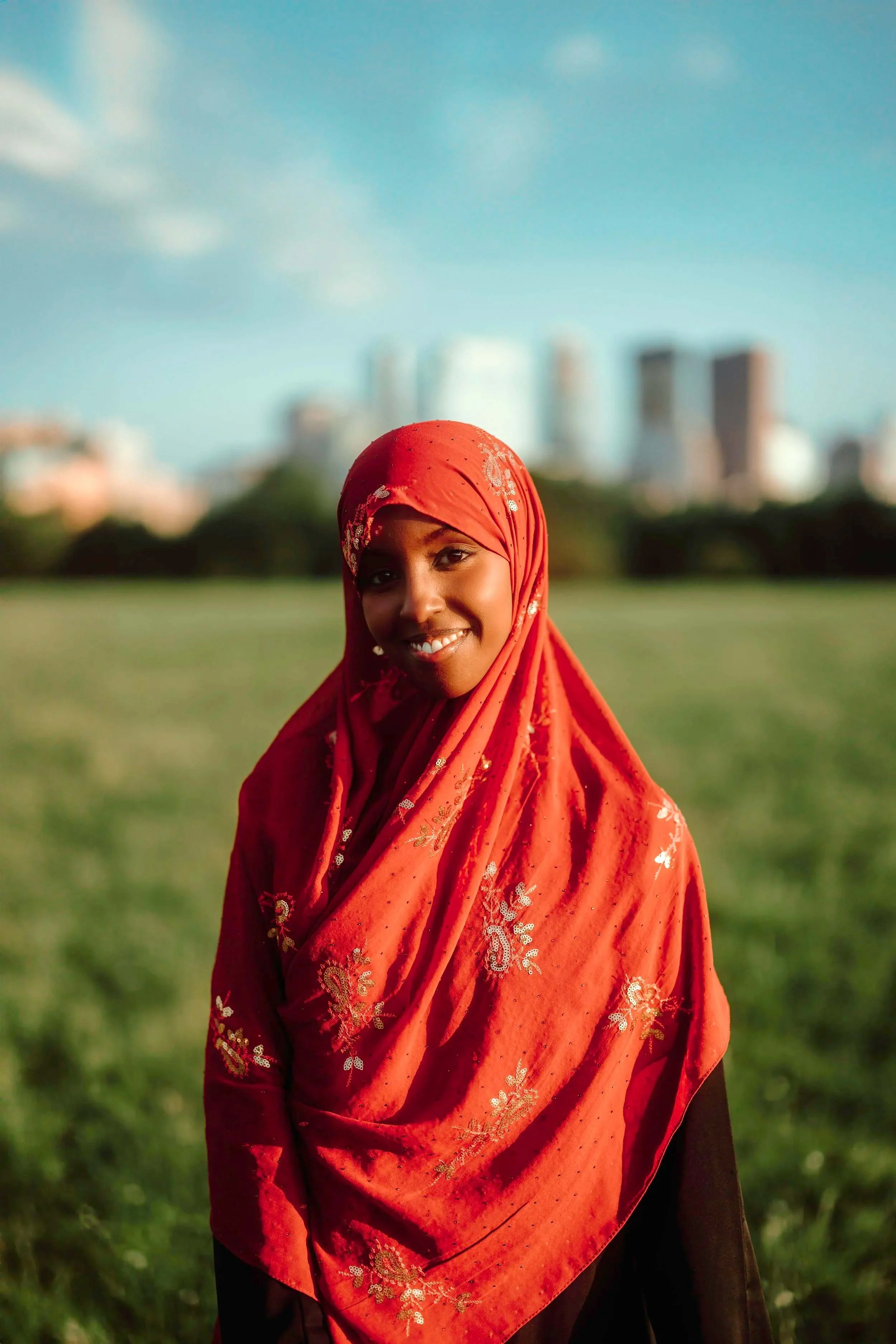 A woman wearing a red, embroidered headscarf stands outdoors on a sunny day, with a grassy park and blurred city skyline in the background.