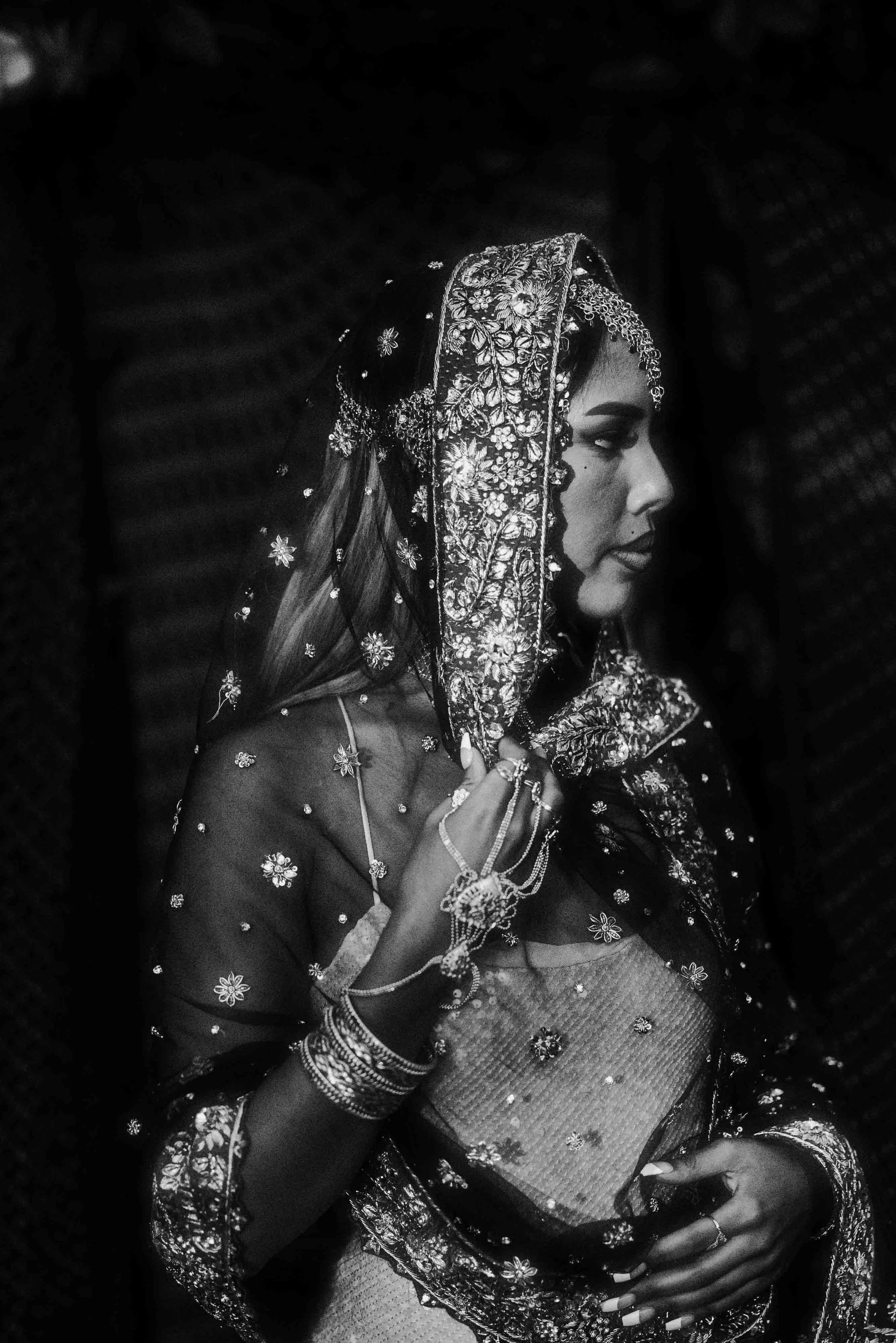 A woman dressed in traditional Indian bridal attire, adorned with intricate jewelry and a heavily embroidered dupatta over her head, is captured in profile in this black and white photo.