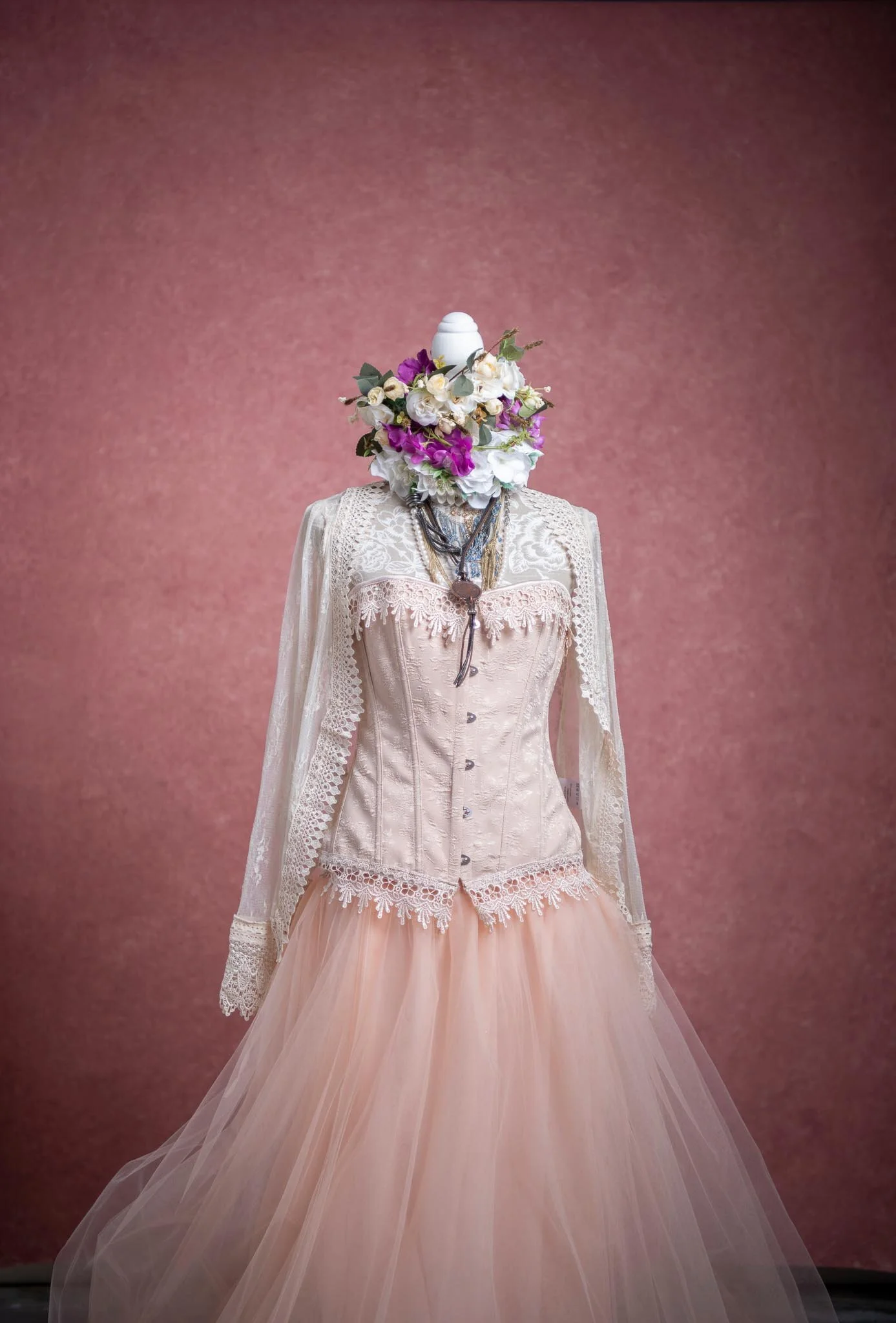 A mannequin dressed in a vintage-style beige corset and peach tulle skirt, topped with an elaborate floral mask made of white, purple, and pink flowers, with a pink background.