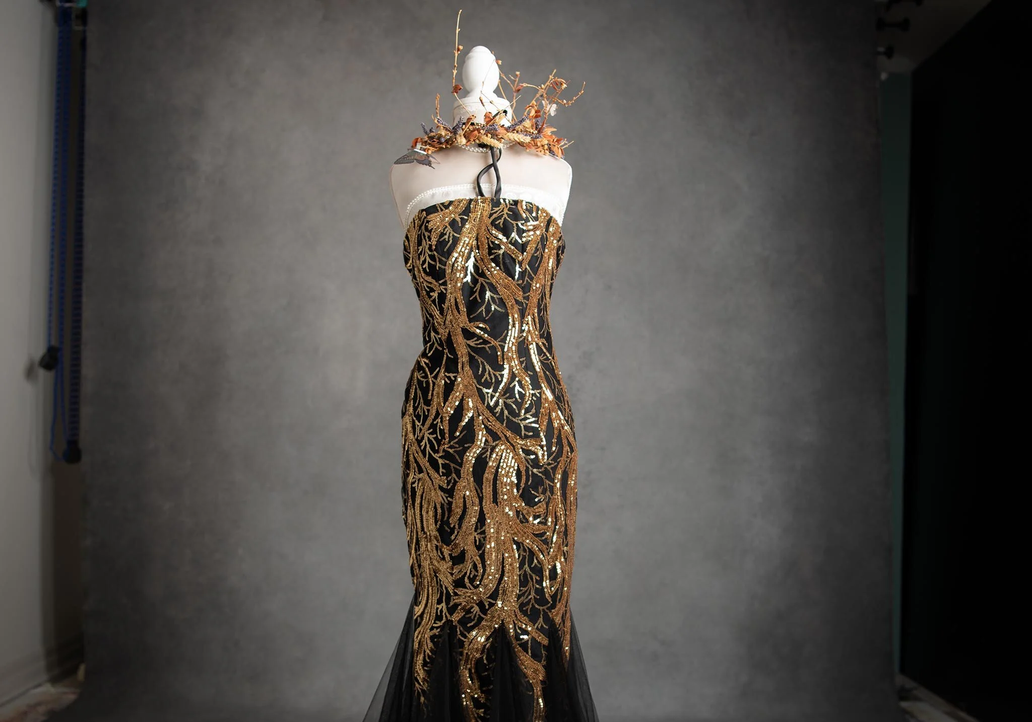 A strapless black gown with intricate gold embroidery and sequin details displayed on a mannequin, with floral embellishments around the neck and shoulders against a plain grey backdrop.