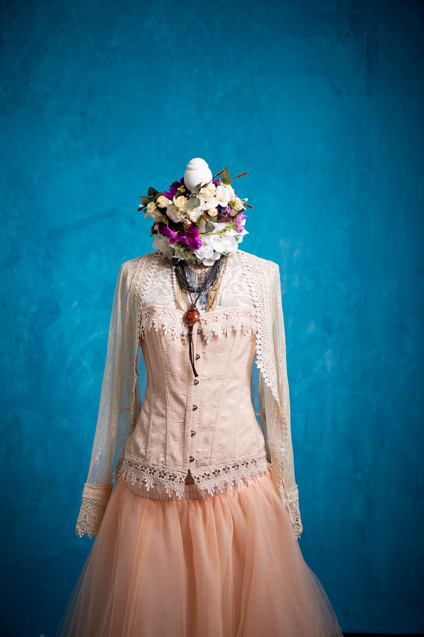 A mannequin dressed in a vintage-style wedding gown with floral accents and layered necklaces, with a head made of white fruit, roses, and purple flowers, against a blue background.