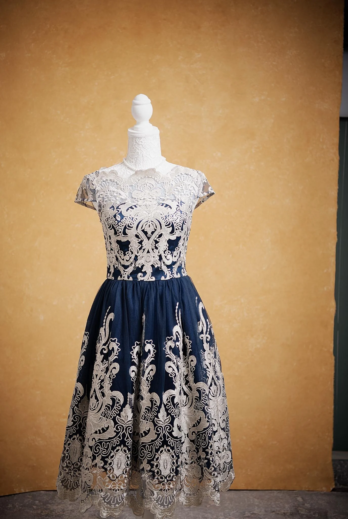 A dress displayed on a mannequin against a yellow background. The dress features intricate white lace details over a dark fabric, with a fitted bodice and a full-length skirt.