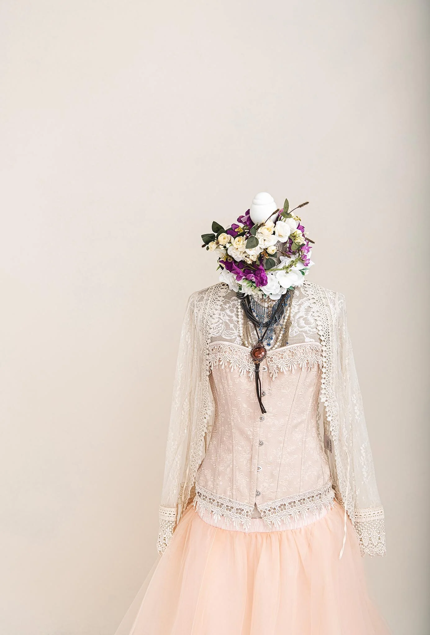 A mannequin dressed in a vintage-inspired wedding gown with lace details, layered skirt, and layered necklaces, with a floral headpiece made of flowers and greenery covering the head area, against a plain beige background.