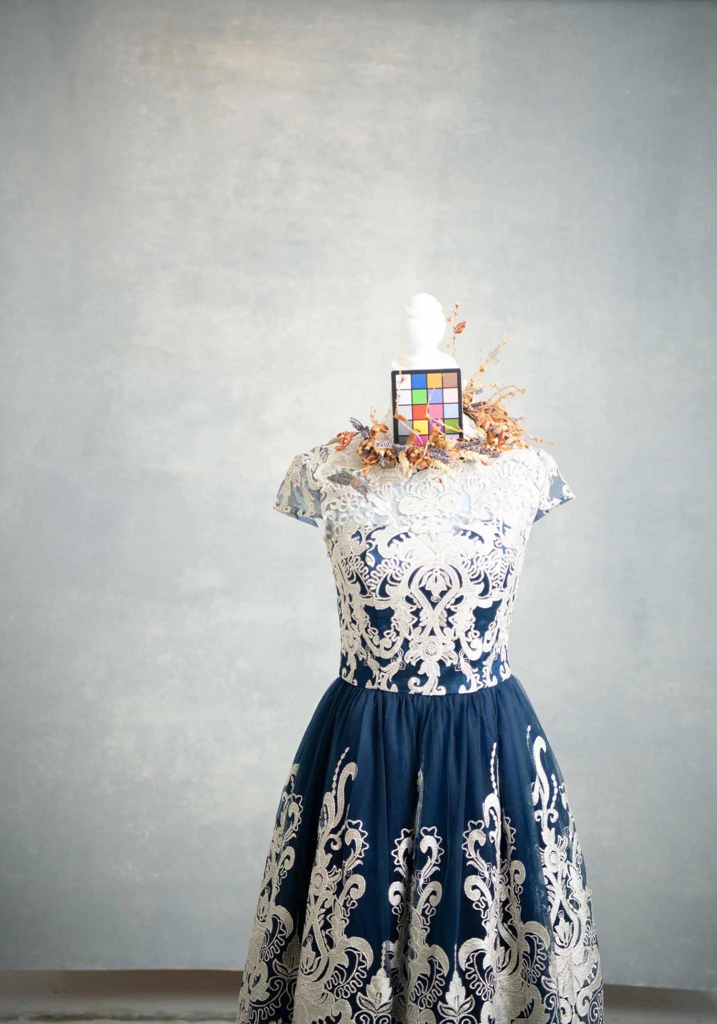 A dress form wearing an intricate navy and white dress with embroidery, topped with a head sculpture, a color calibration chart, and decorative dried leaves.